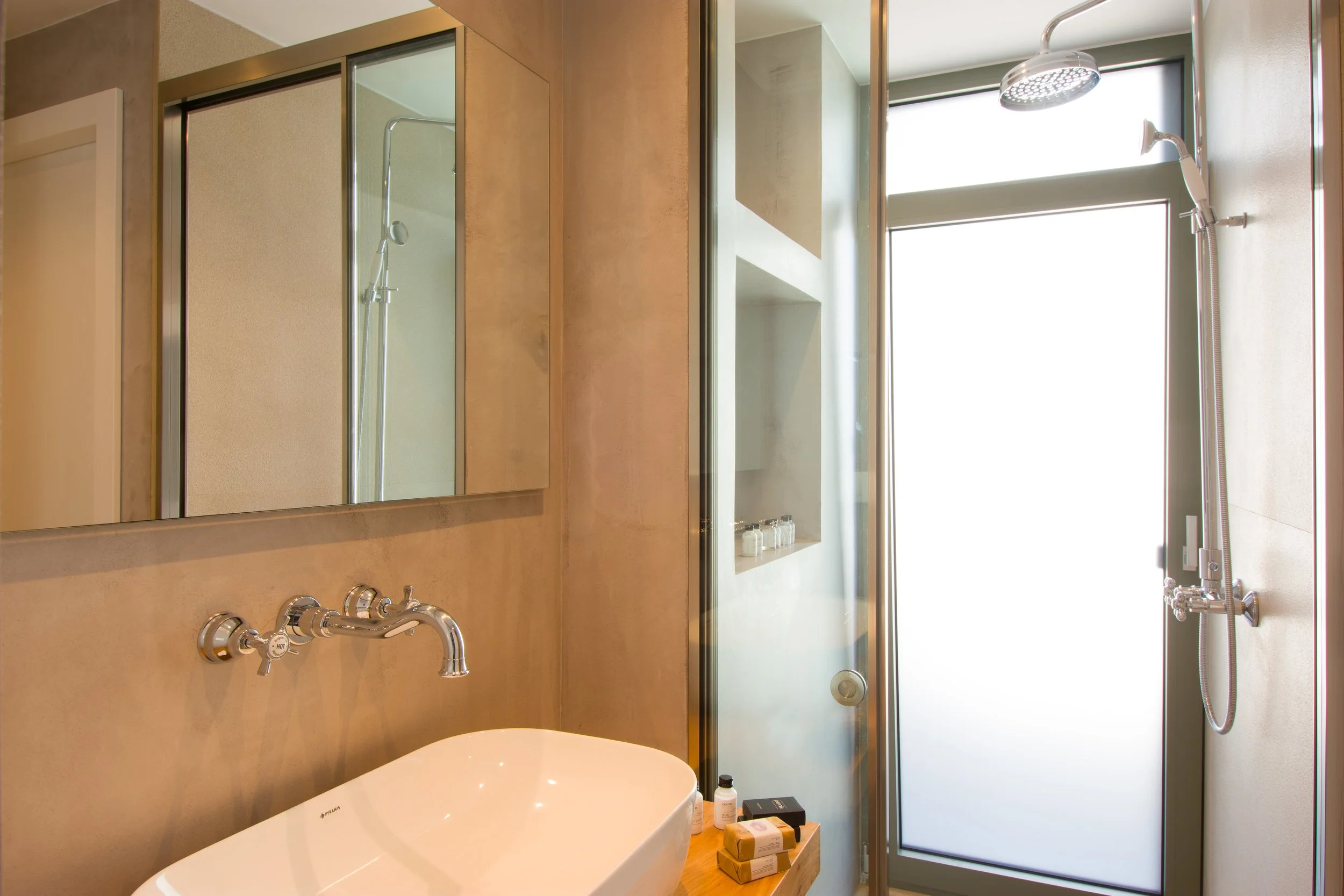 Modern bathroom features a wall-mounted sink, a large mirror, a glass shower enclosure, a handheld showerhead, and shelves with toiletries.