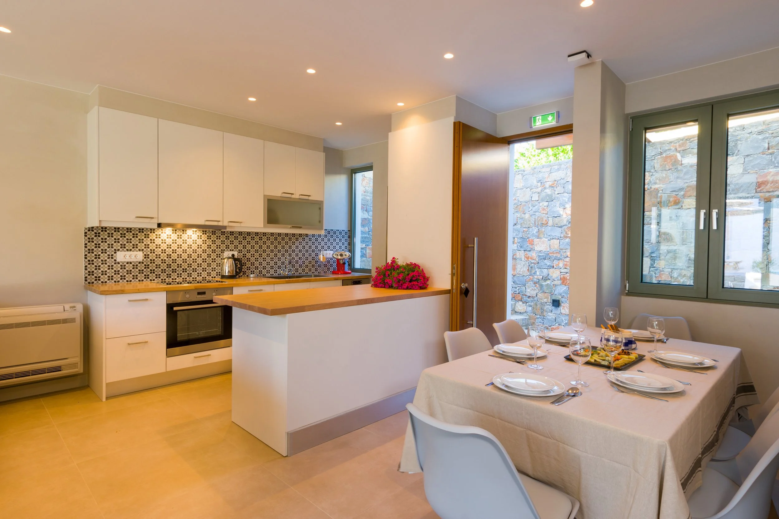 Modern kitchen and dining area with a table set for a meal, featuring white chairs, a white tablecloth, wine glasses, and a platter of food, with natural light coming through large windows and a stone wall outside.