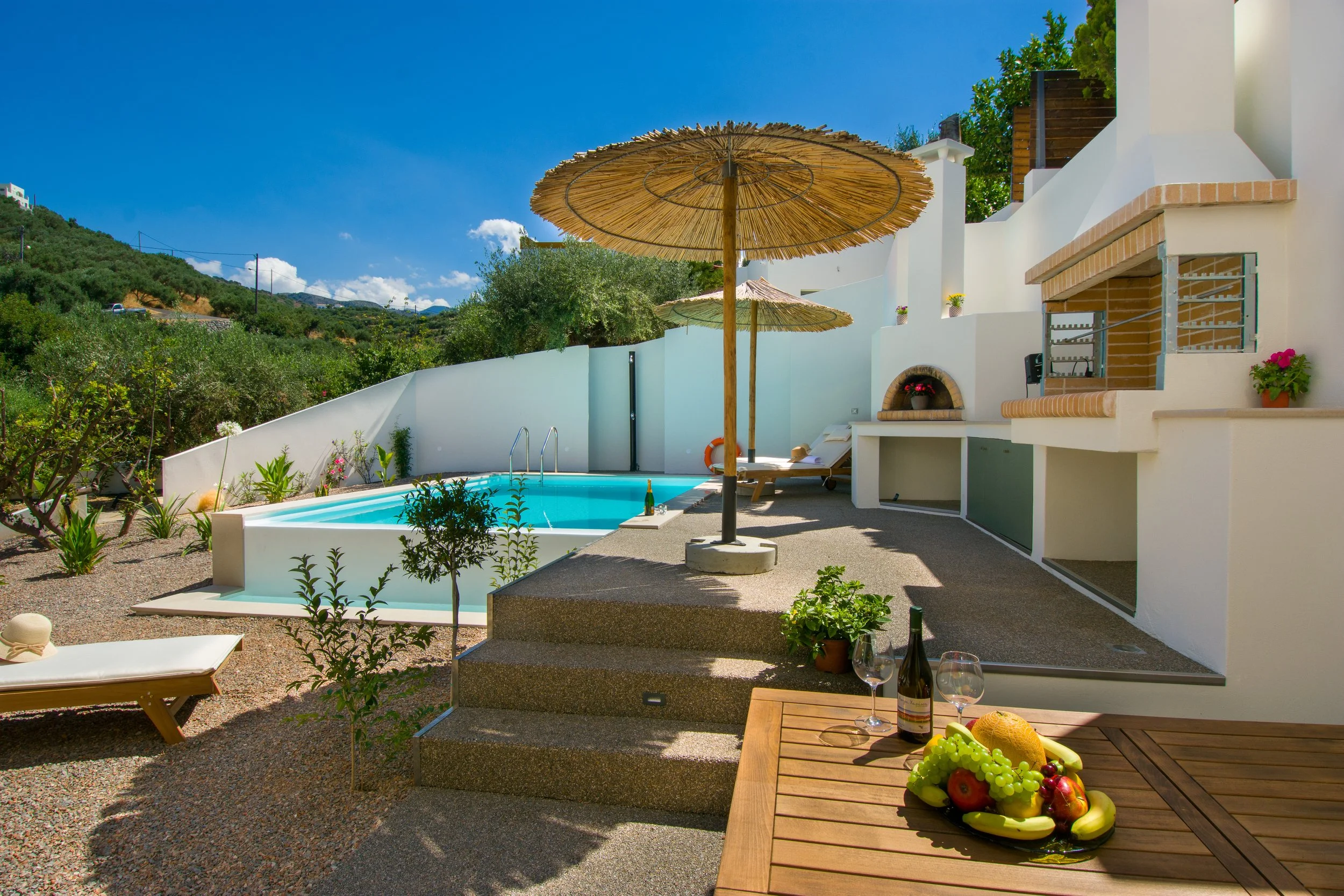 A sunny outdoor pool area with two straw umbrellas, lounge chairs, surrounding greenery, a white house with a small fireplace decorated with flowers, and a table with grapes, apples, and a bottle of wine.