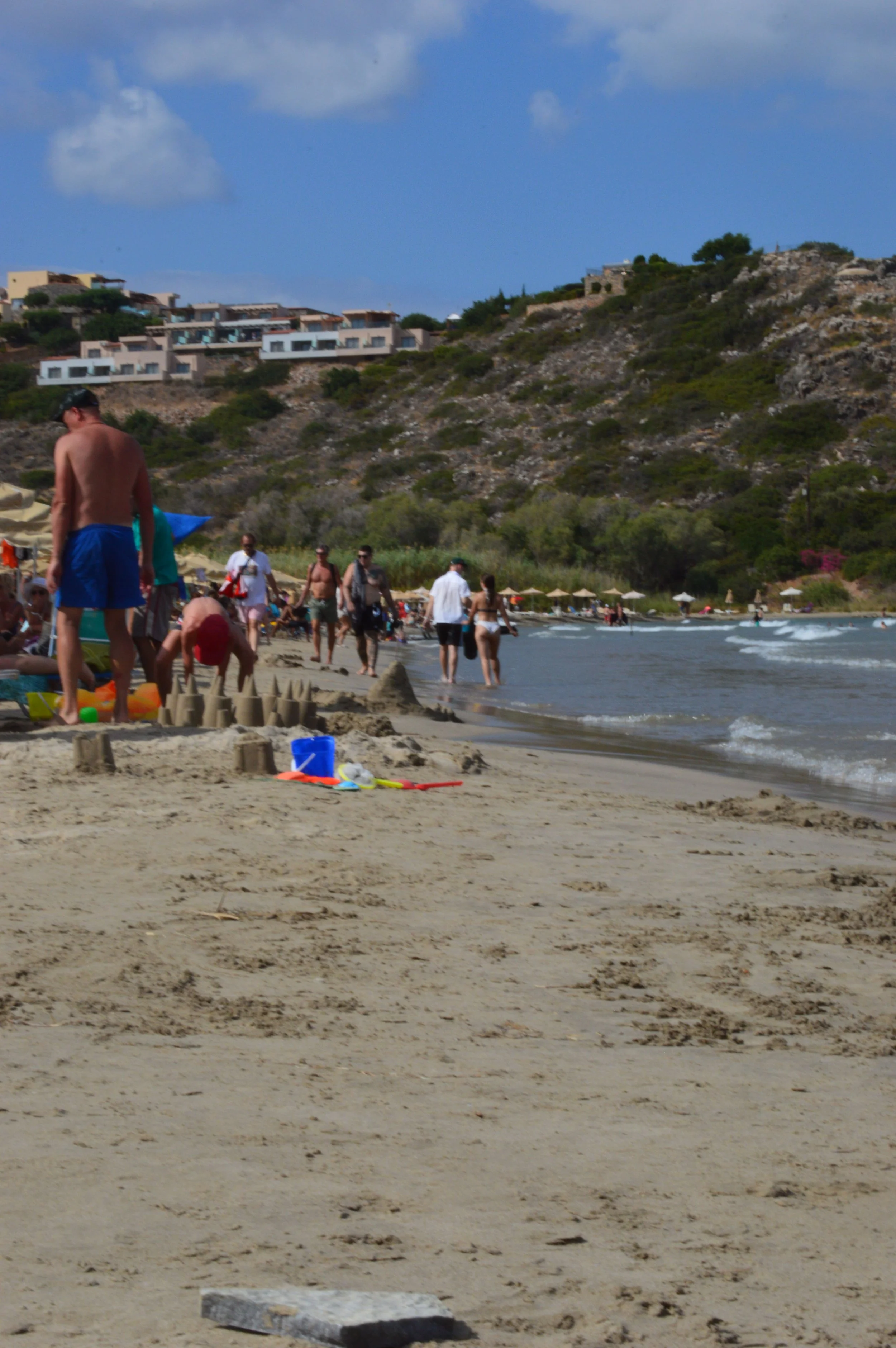 People walking along a sandy beach with umbrellas and sandcastles, hillside with modern houses and rocks, blue sky with clouds.