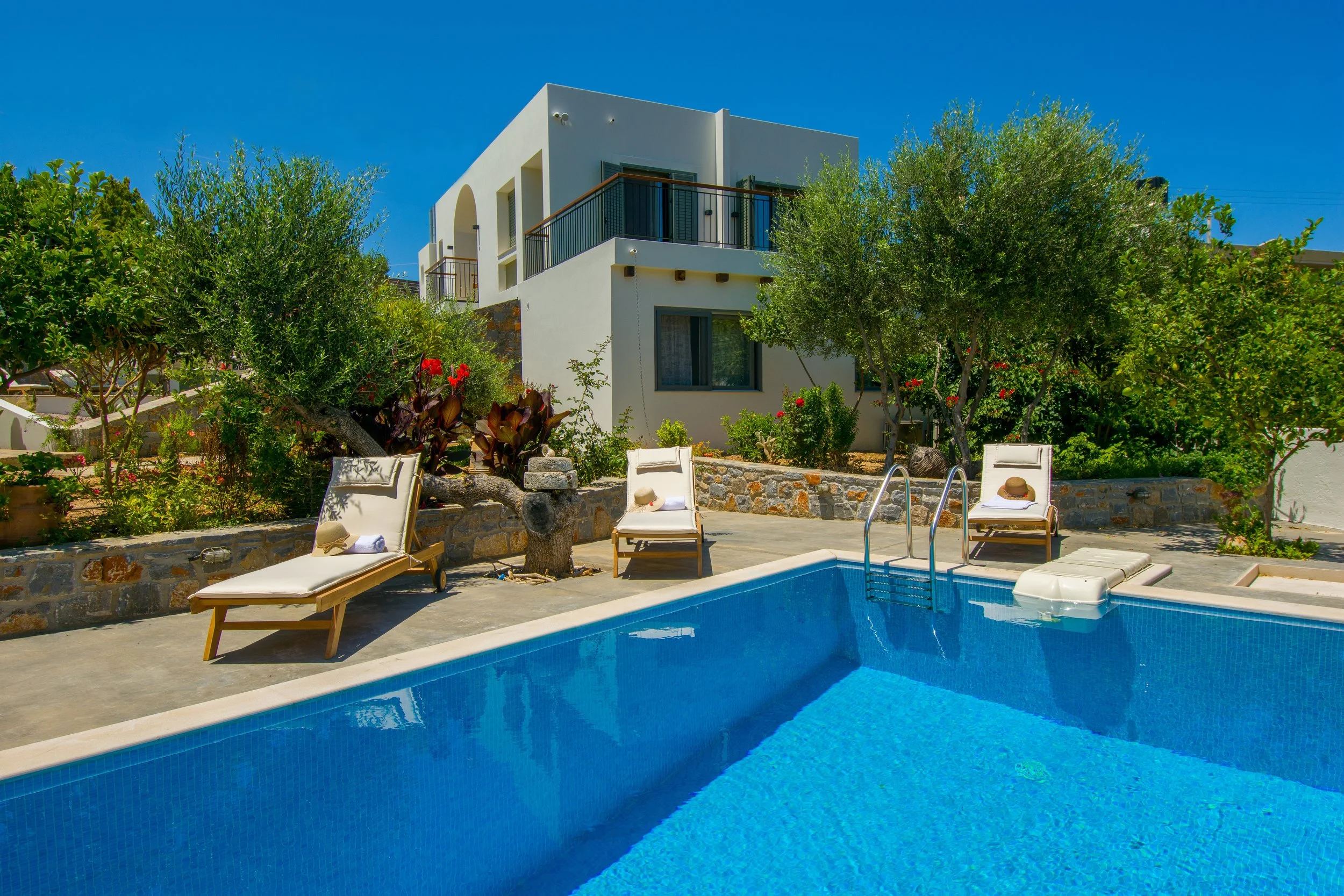 A sunny backyard swimming pool with blue water, surrounded by lounge chairs, a hat, and towels, with a modern white house and lush greenery in the background.