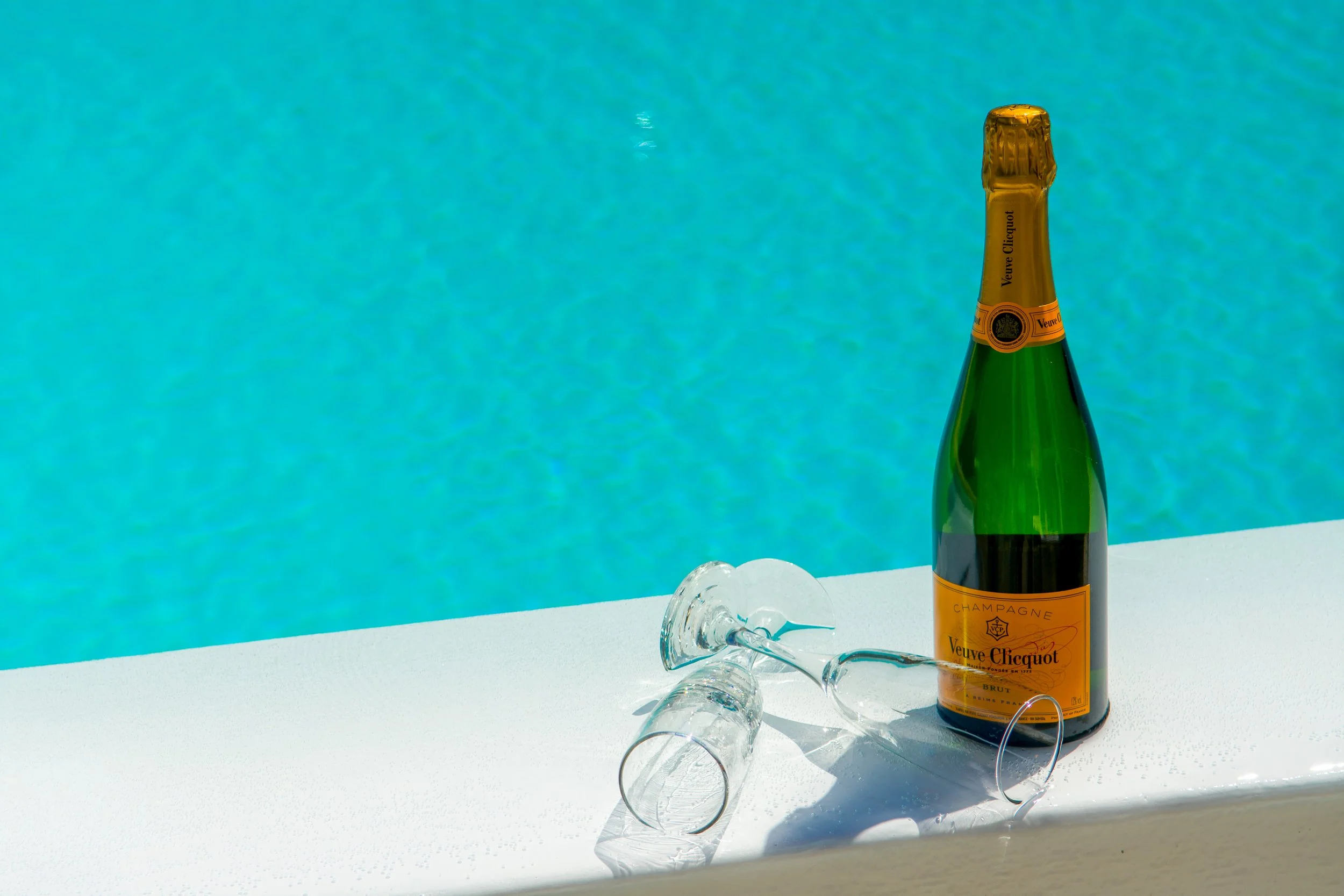 A bottle of Veuve Clicquot champagne with a golden foil top, a signed label, and a glass stem in front of a turquoise background, with ice scattered on the white surface.