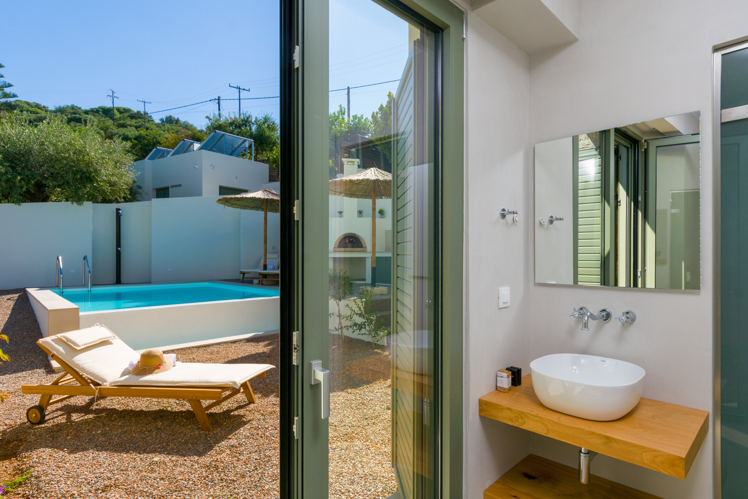 Interior view showing a bathroom with a white round bathtub and a wooden countertop, looking out to an outdoor pool area with a lounge chair, a straw hat, and a pool with clear blue water, surrounded by a white wall, umbrellas, and hills with trees a