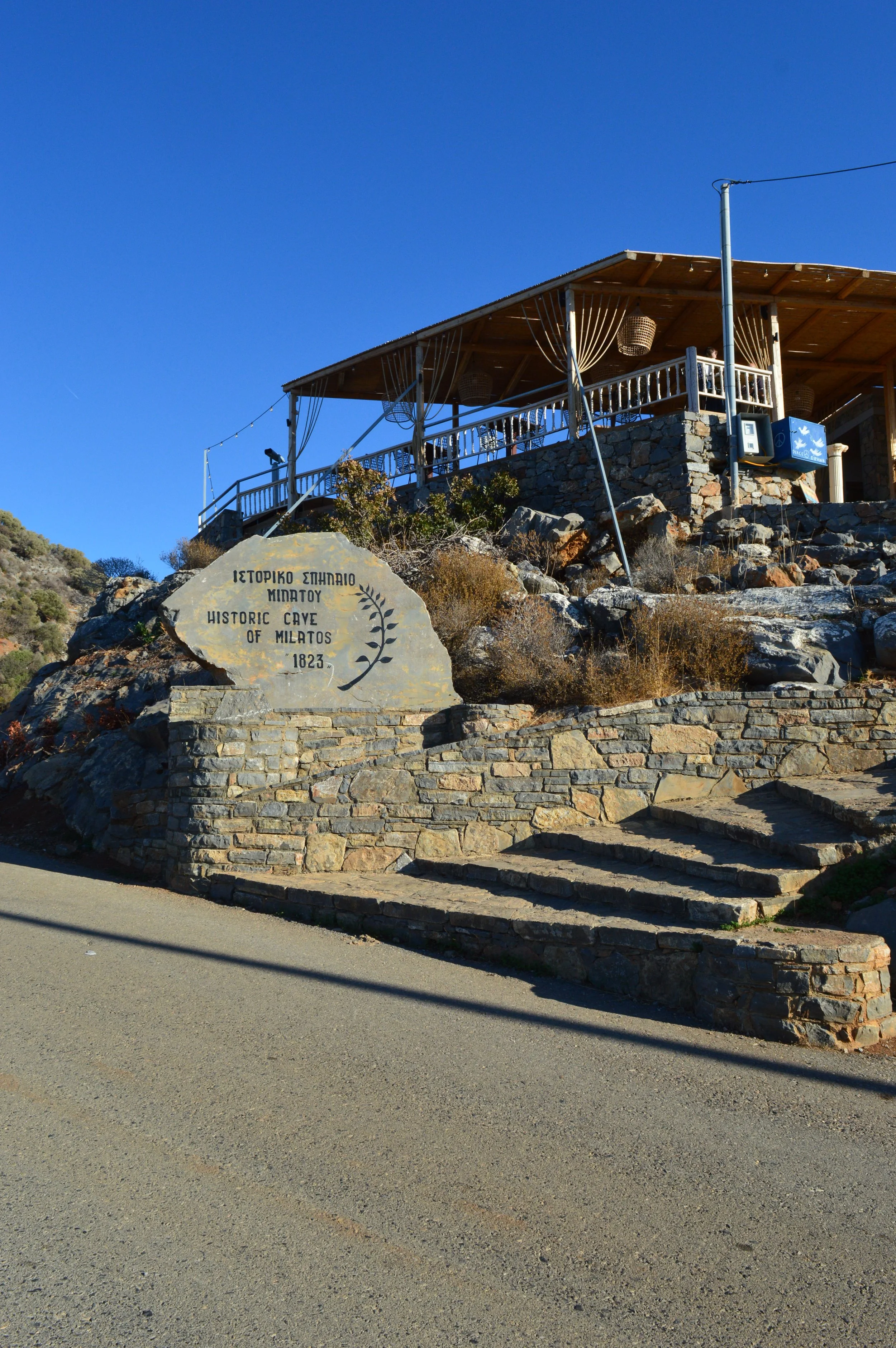 A stone sign with Greek and English inscriptions reading 'Historic Cave of Milatos, 1823' placed on a rocky slope. A wooden structure, possibly a café or lookout, is built on the elevated part of the rocky terrain with a railing and hanging lanterns. There are stairs and a paved path leading up to it, with clear blue sky in the background.