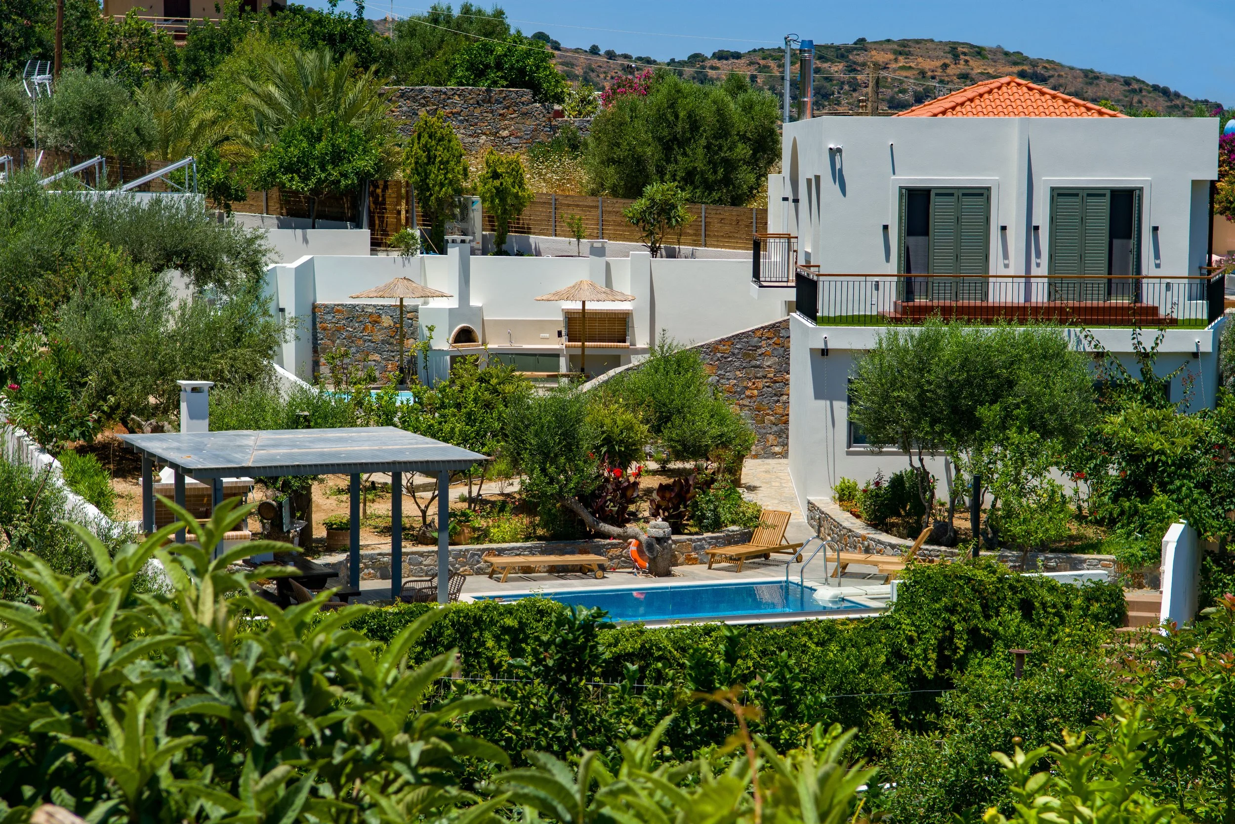 A hillside residential area with white modern houses, surrounded by lush greenery and trees. There is a swimming pool with lounging chairs and umbrellas, and mountains are visible in the background under a clear blue sky.