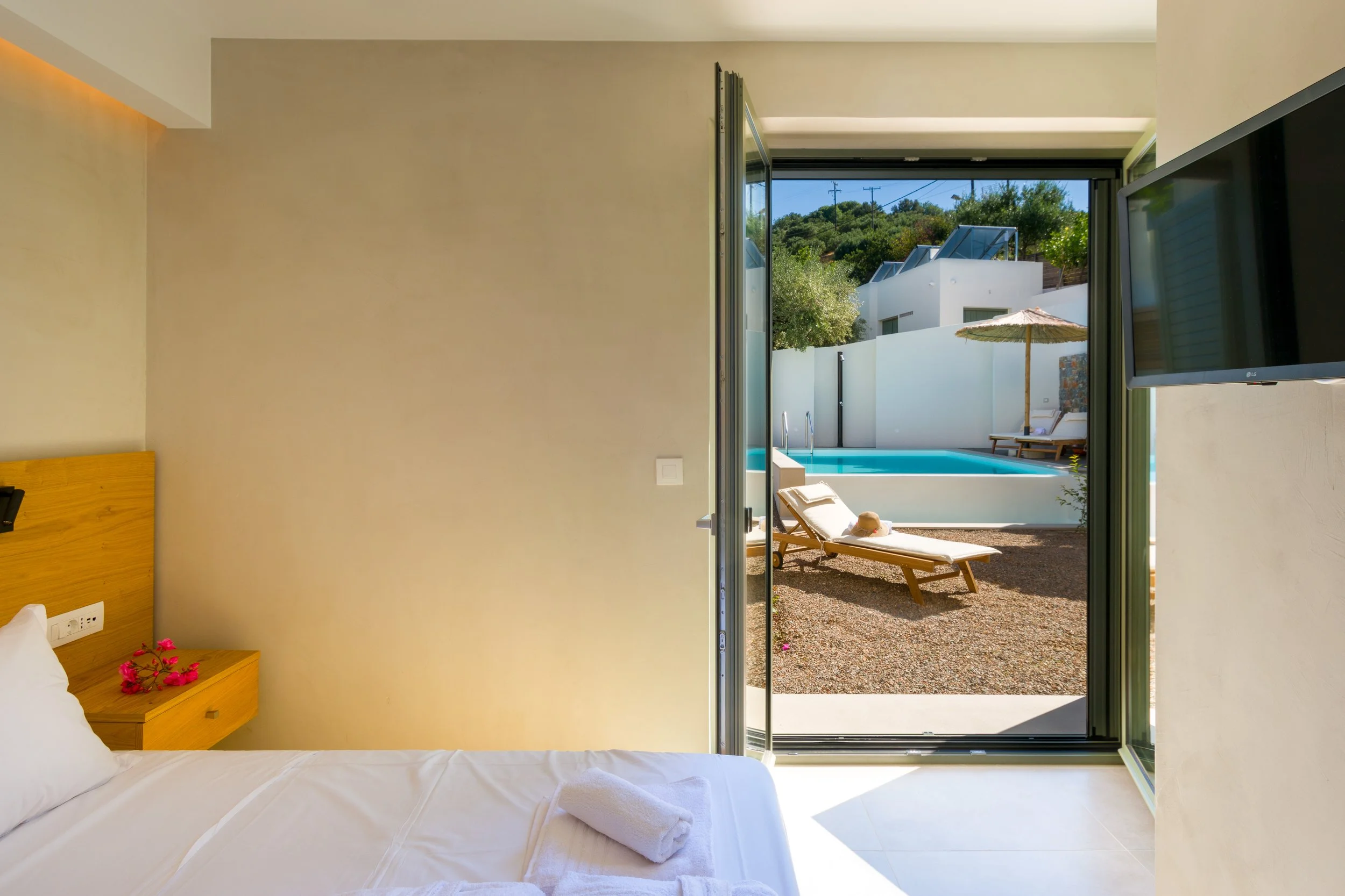 Milatos Garden Villas - A hotel room with a bed, bedside table with pink flowers, and an open door leading to a patio with a swimming pool, lounge chairs, an umbrella, and a view of neighboring buildings and greenery.