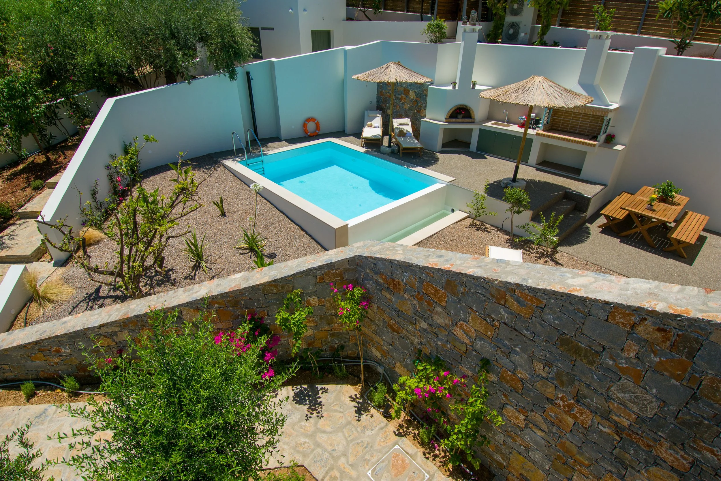 Milatos Garden Villas - A backyard swimming pool with lounge chairs and thatched umbrellas, surrounded by a white boundary wall, gardens, and trees.