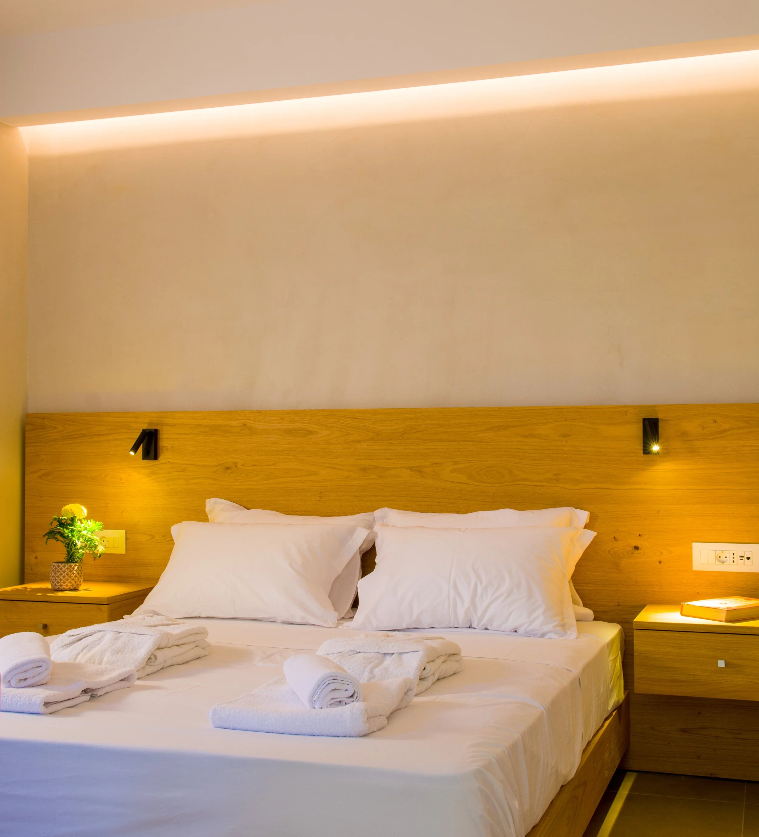 A neatly made bed with white linens and pillows, with rolled-up towels on top. The bed has a wooden headboard with two small black reading lights above it, and a nightstand with a potted plant next to the bed. The wall behind the bed is painted light