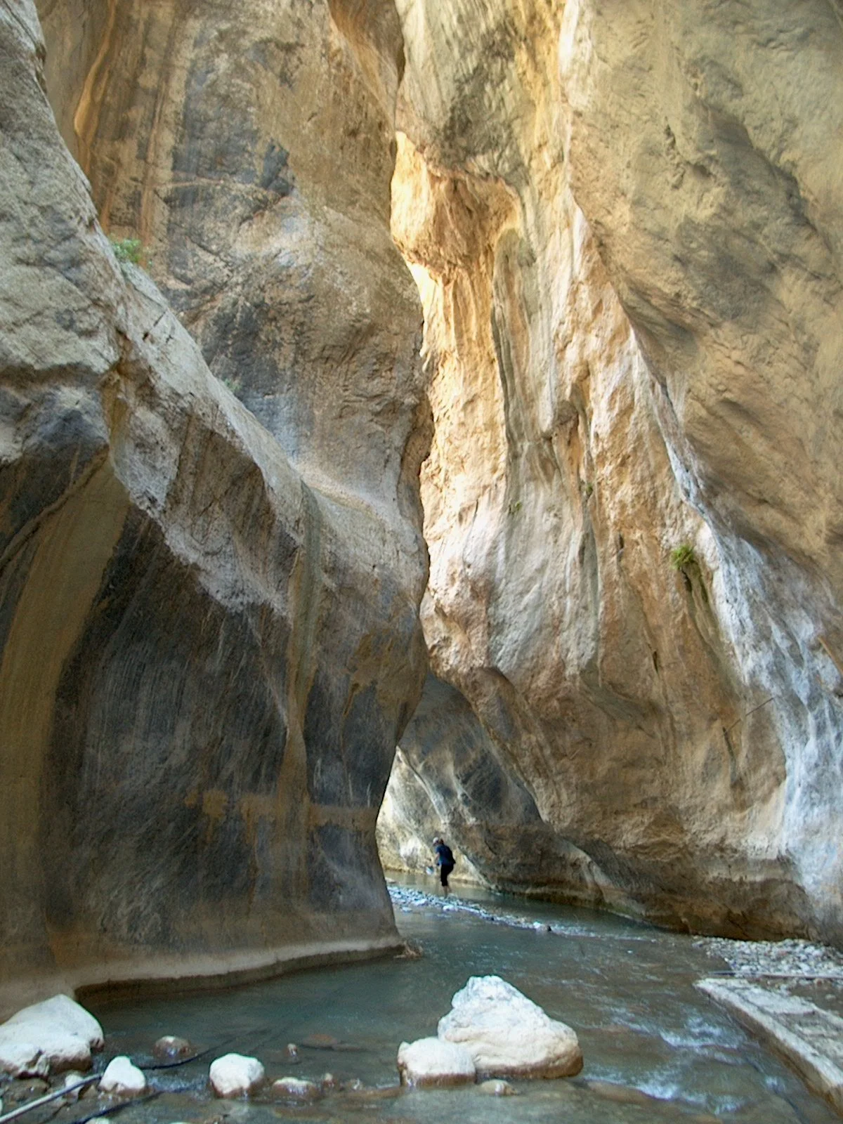 Milatos Garden Villas - Sarakina gorge - A narrow canyon with tall, layered rock walls and a small stream flowing through the canyon floor. A person is seen walking in the distance.