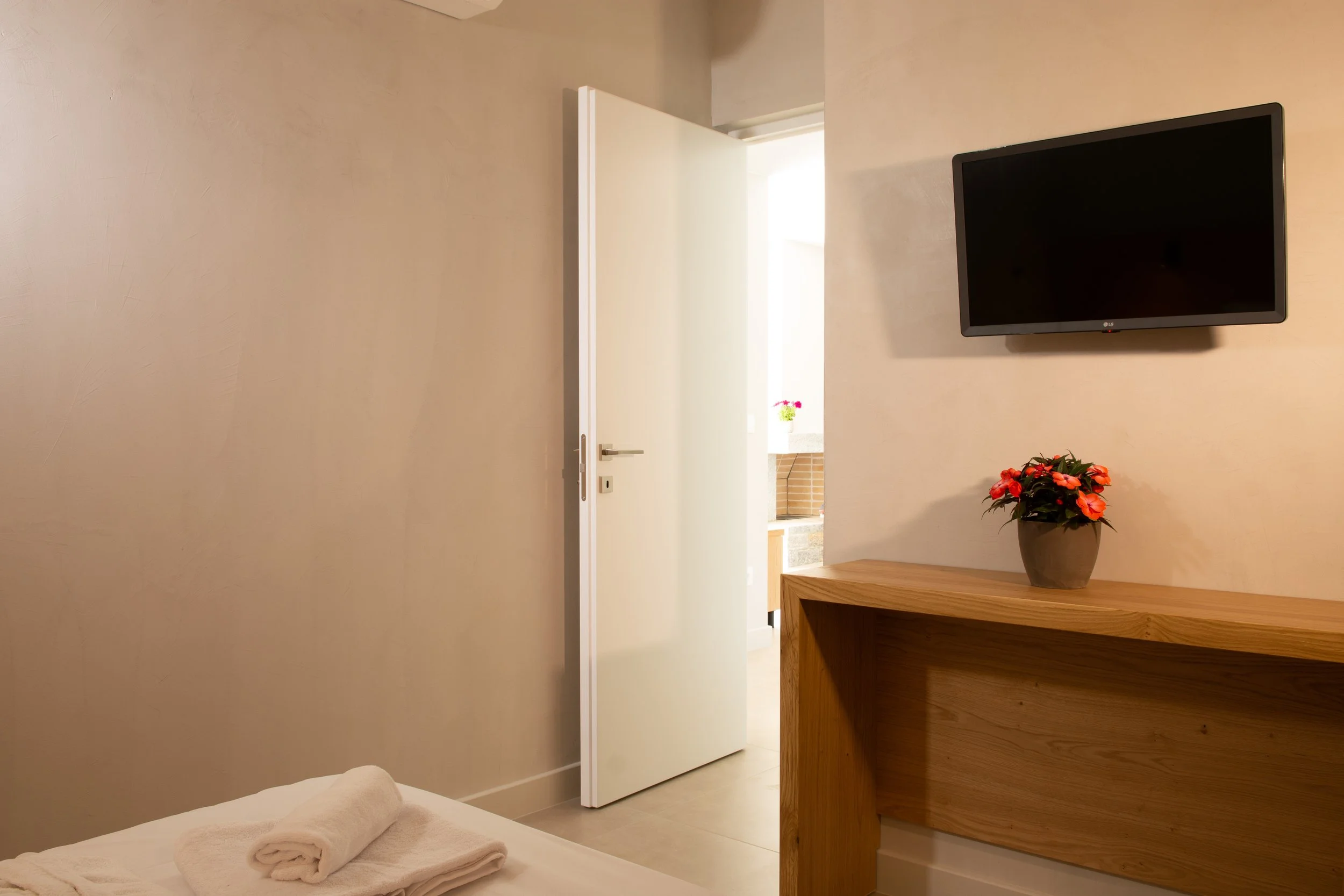 A hotel room with a bed, folded towels, a small wooden table with a potted plant with red flowers, a mounted flat-screen television, and an open white door leading to another room or balcony.