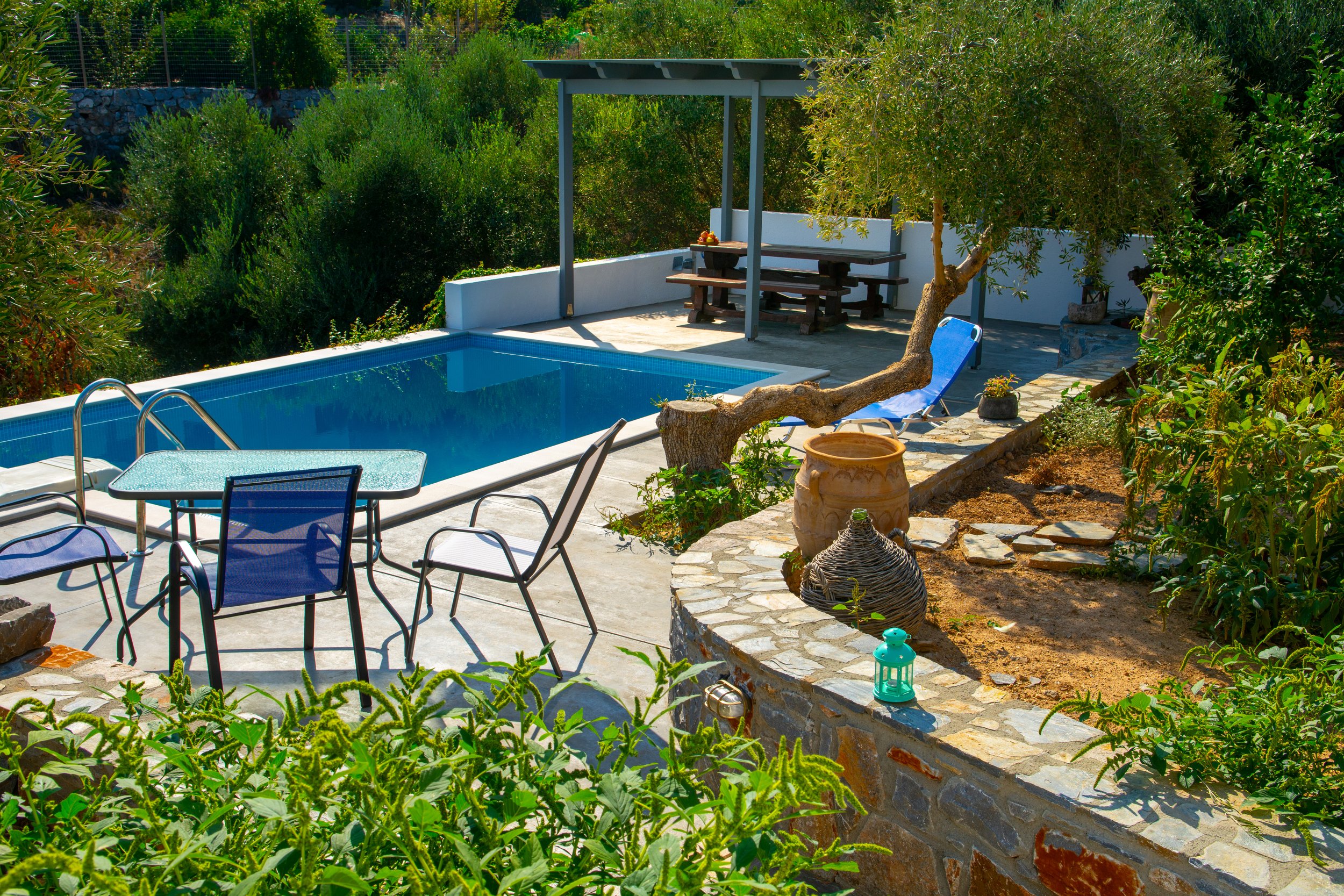 A backyard with a swimming pool, green bushes, outdoor seating, a tree, potted plants, and a shaded dining area with a picnic table.