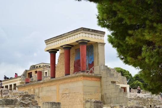 Ancient Greek ruins with red columns and a partially restored building, surrounded by trees and cloudy sky.