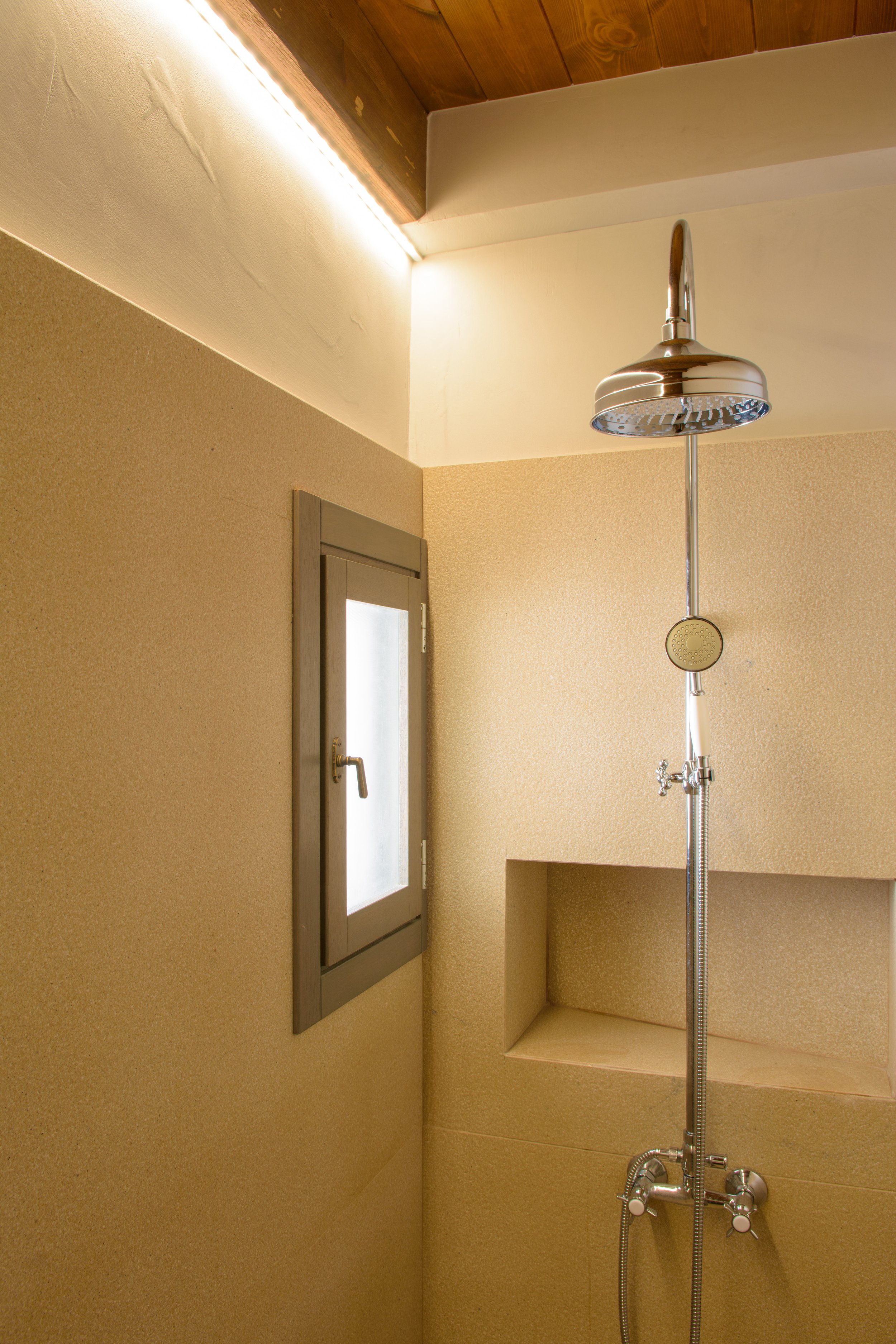 A shower area with a chrome showerhead attached to a flexible hose, mounted on a wall with beige textured wallpaper. There is a small, rectangular mirror with a gray frame and a frosted glass door with a handle on the left side. The ceiling has wood 