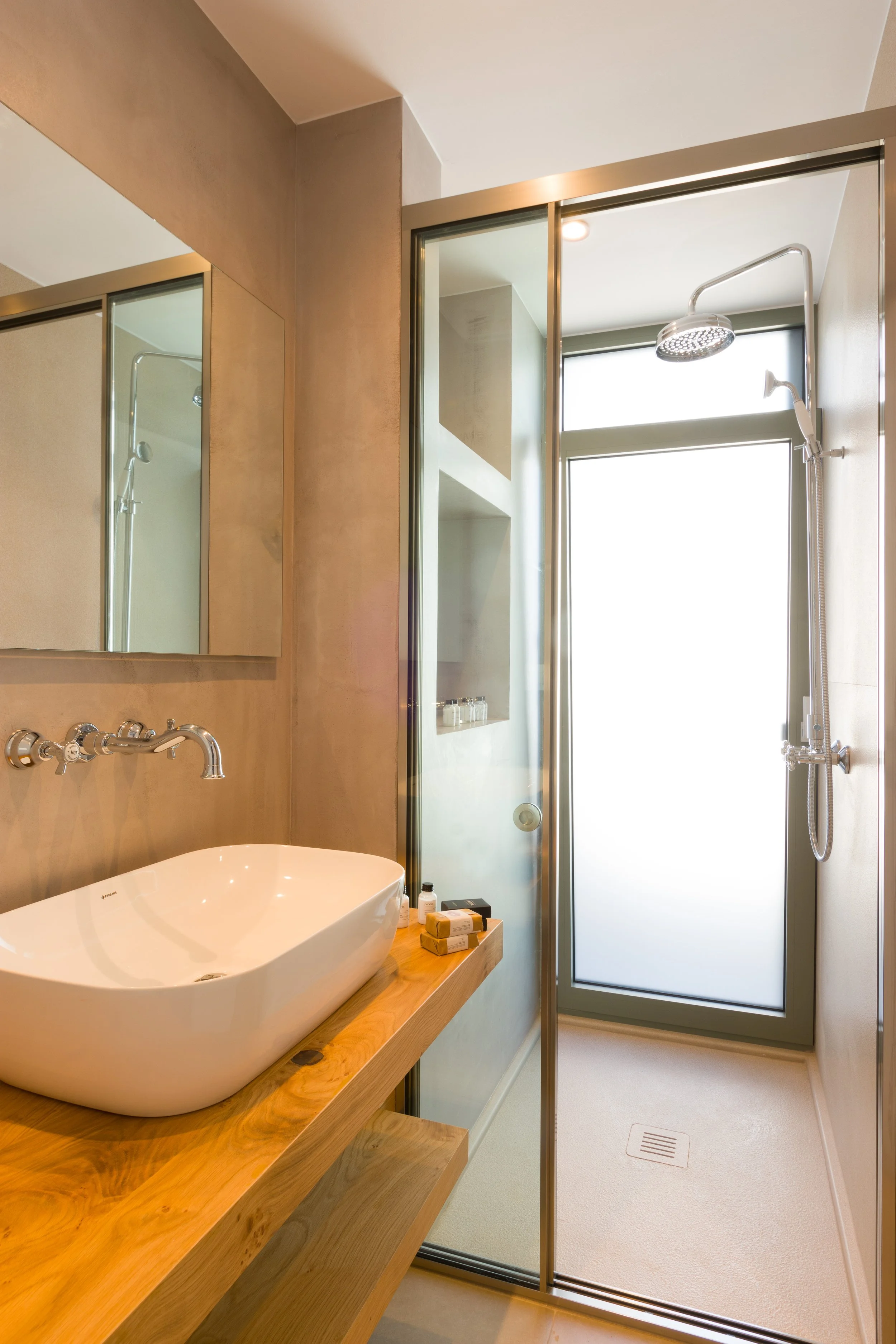 A modern bathroom with a wooden floating vanity holding a white vessel sink and bottles of toiletries. There are beige walls, a large mirror, and a frosted glass shower enclosure with a sliding door. Inside the shower, a rain showerhead and a handhel