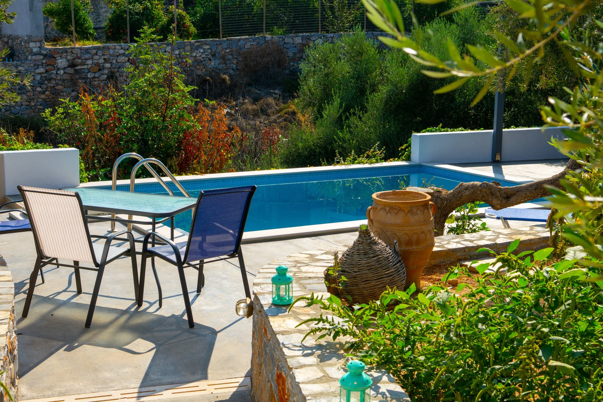 A backyard with a small rectangular swimming pool, surrounded by outdoor furniture including chairs and a table. There are decorative plants and a stone wall in the background, with lush greenery and trees providing cover.