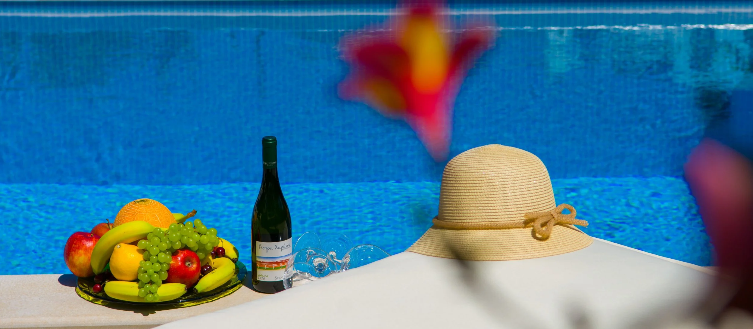 A table with a fruit platter, a bottle of wine, empty wine glasses, a straw sun hat, and a person holding a red and yellow flower in front of a swimming pool.