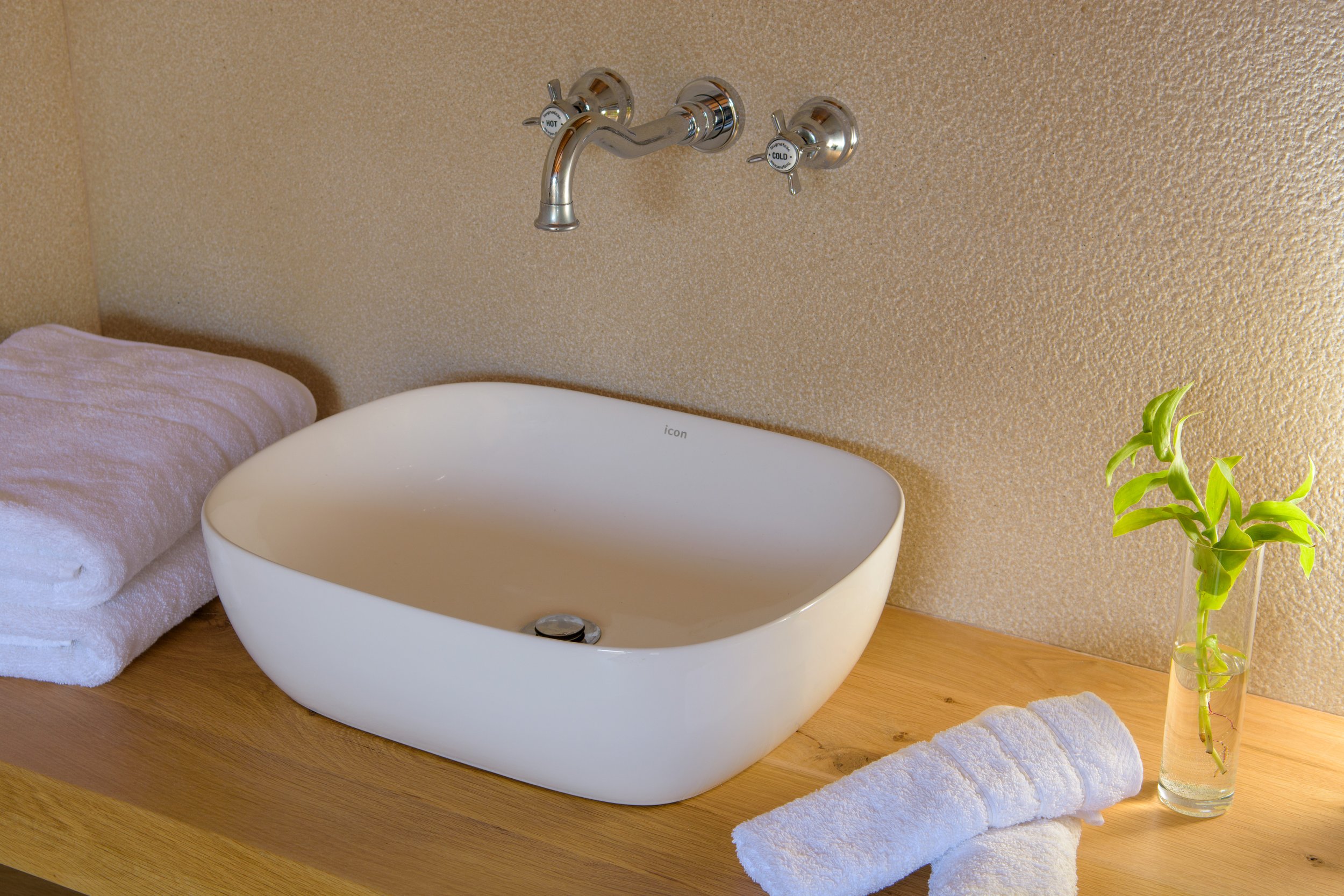 Bathroom sink with a modern oval white basin, a two-handle wall-mounted faucet labeled cold and hot, a rolled white towel on a wooden countertop, and a glass vase with a green plant, all against a textured beige wall.