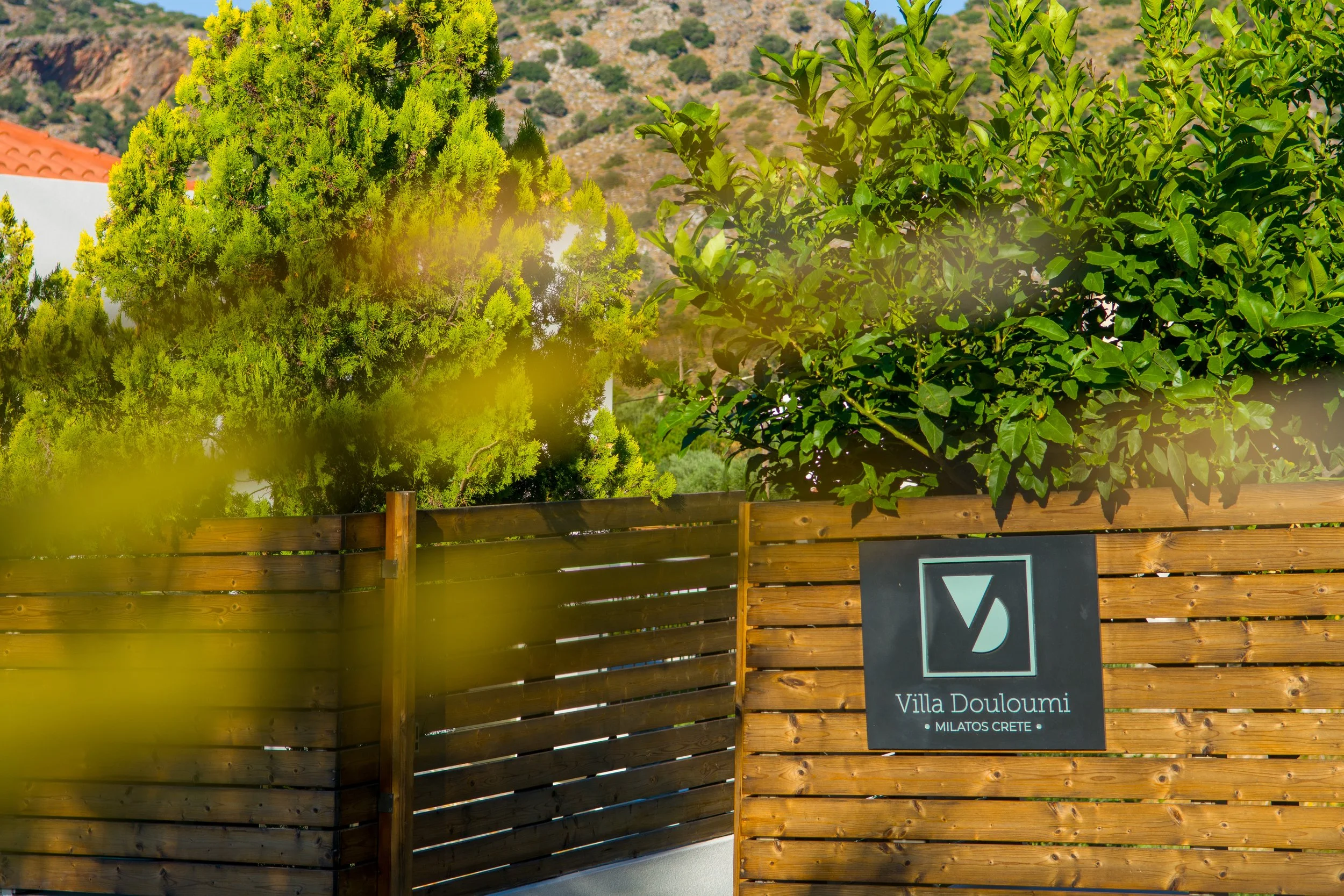 A wooden fence with a sign that reads 'Villa Douloumi, Milatos Crete' in front of green trees and hills during daytime.