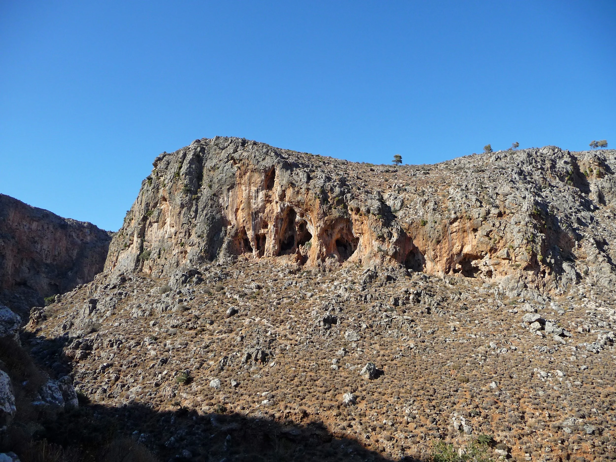A rugged mountain landscape with steep, rocky cliffs and sparse vegetation, under a clear blue sky.
