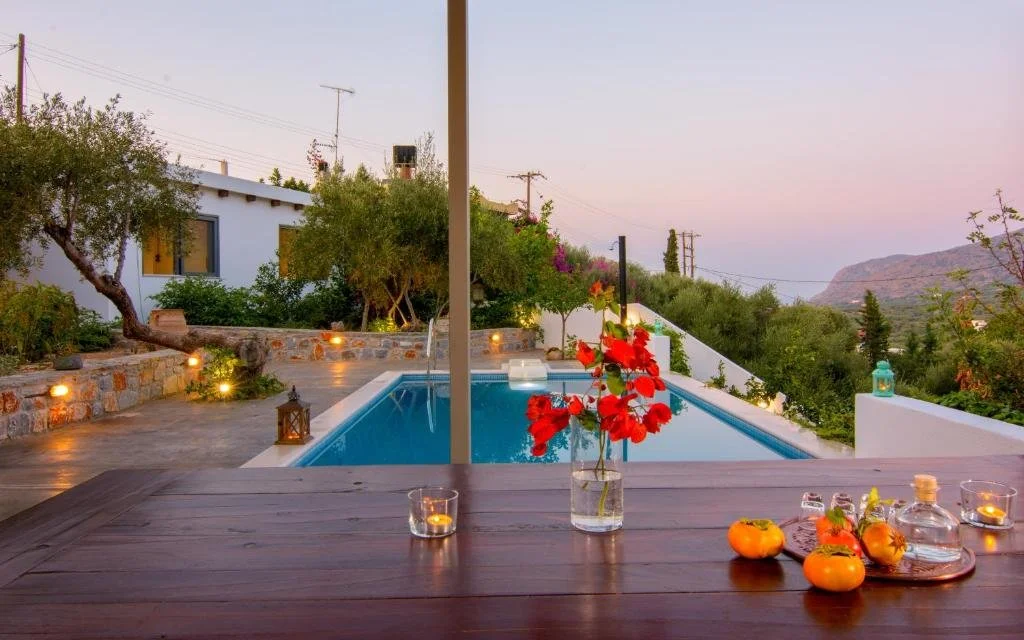 Outdoor swimming pool surrounded by trees and plants, with a wooden table in the foreground holding flowers, candles, and small decorative pumpkins. The scene is set during dusk over a hilly landscape.