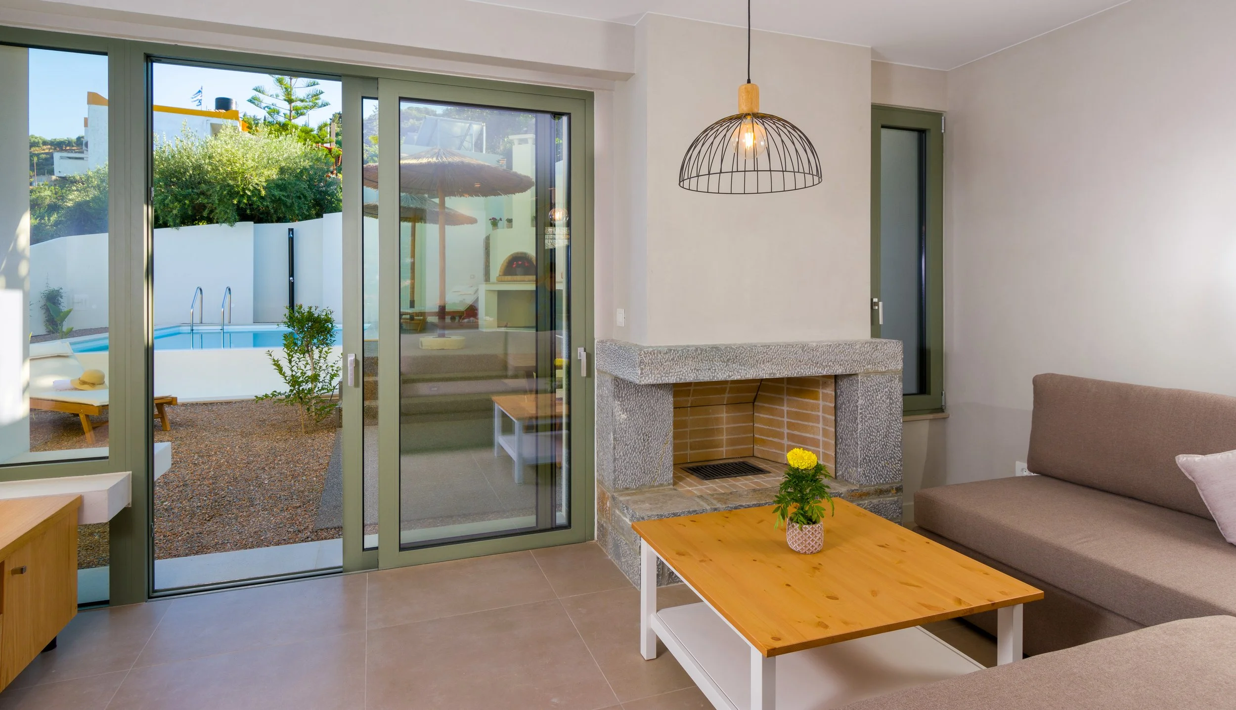 Living room with glass sliding door leading to a pool and outdoor seating area, featuring a beige sofa, wooden coffee table with a small potted plant, a fireplace, and hanging light fixture.