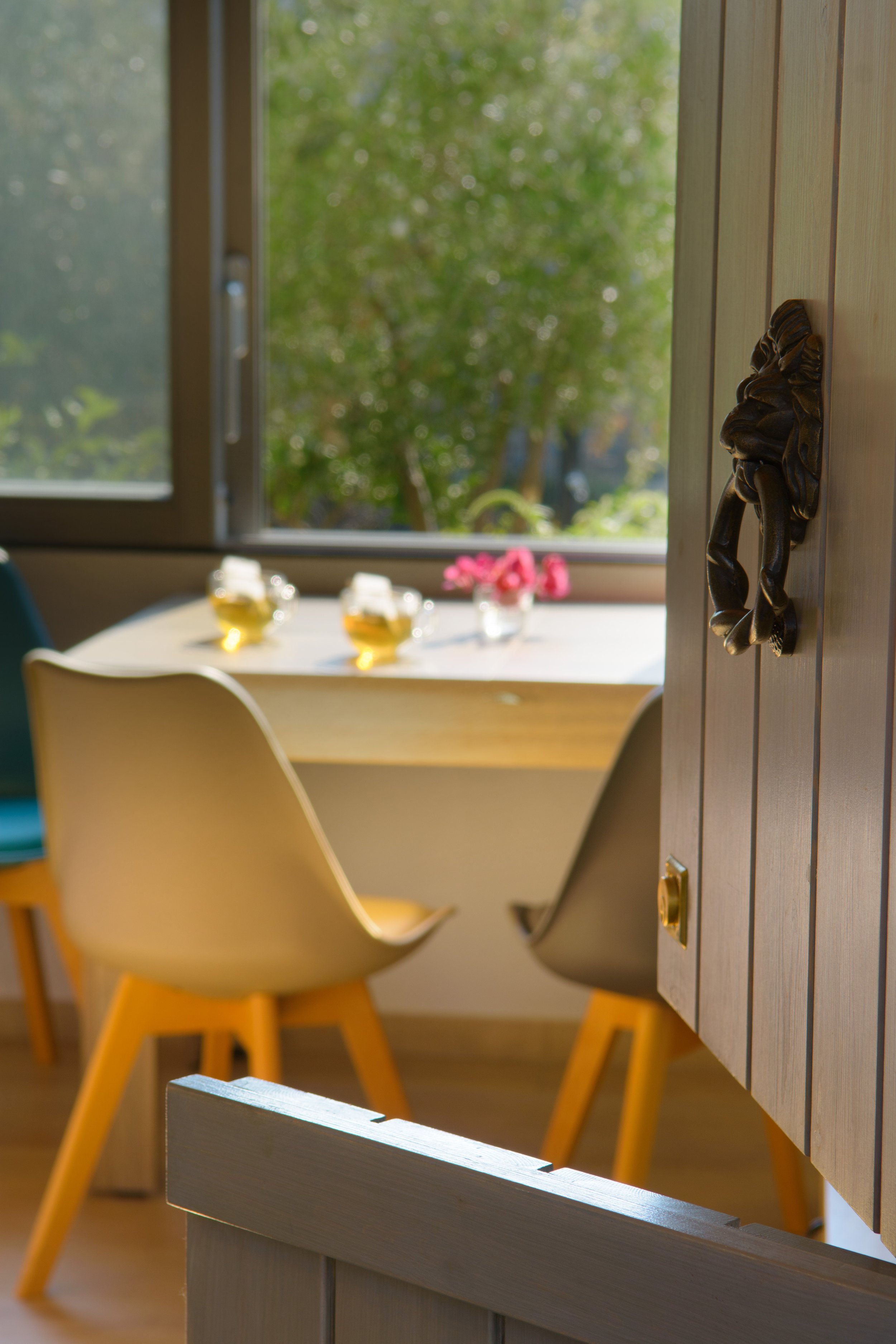 Close-up view of a door handle shaped like a lion's head on a wooden door, with a dining area and two glasses of beer on a table in the background near a window showing trees outside.