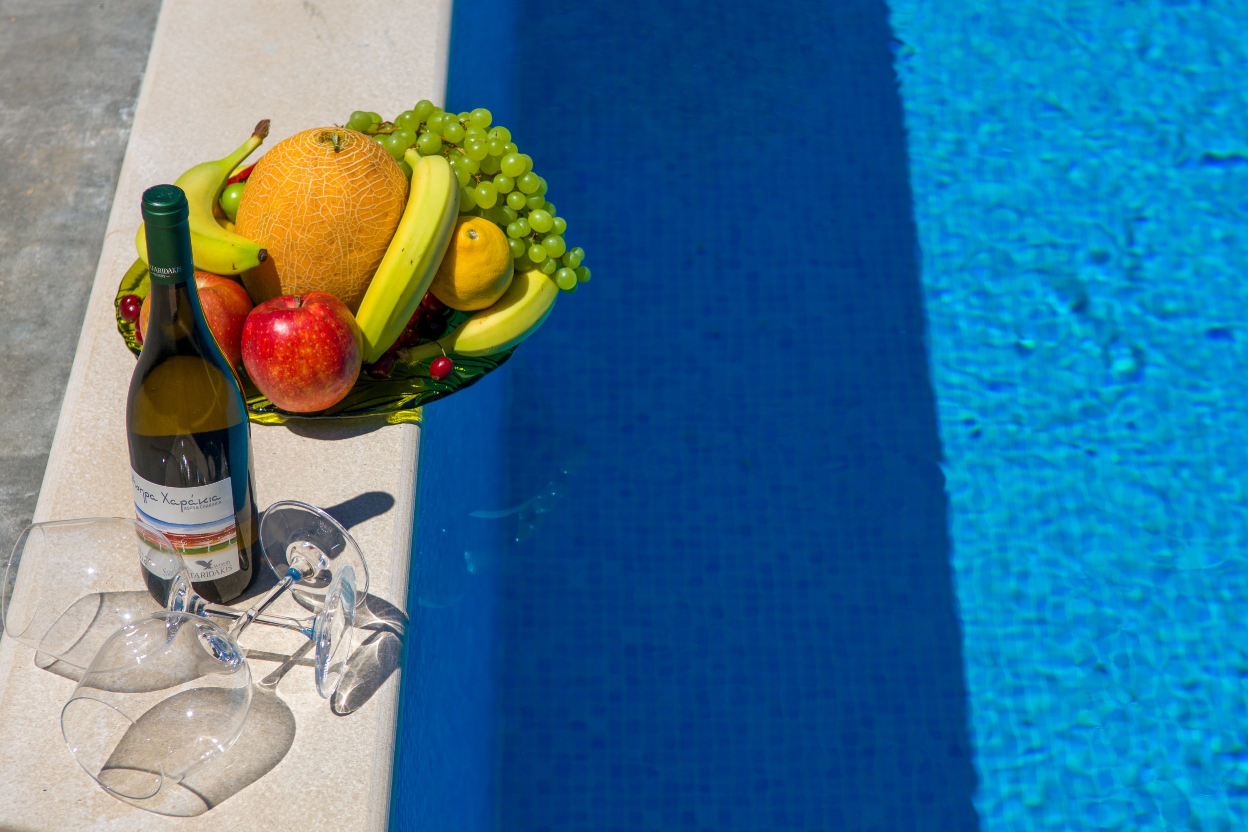 A bowl of fresh fruits, a bottle of wine, and empty wine glasses beside a swimming pool.
