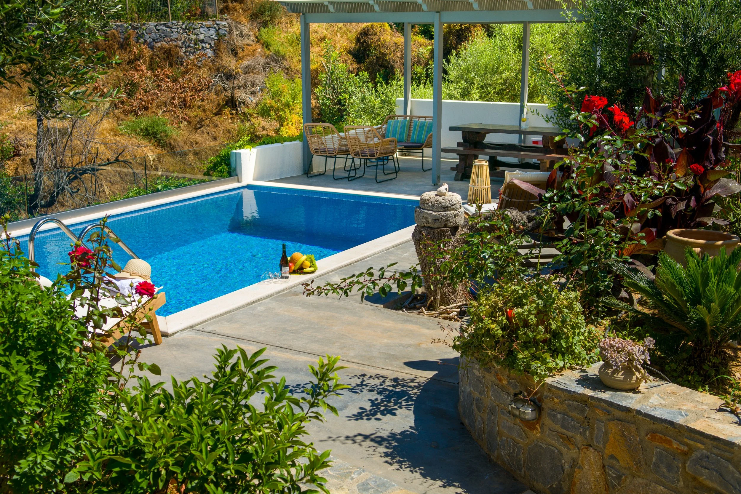 A backyard with a swimming pool, surrounded by various plants and flowers, with outdoor furniture, a wine bottle, and a bowl of fruit on the edge of the pool.