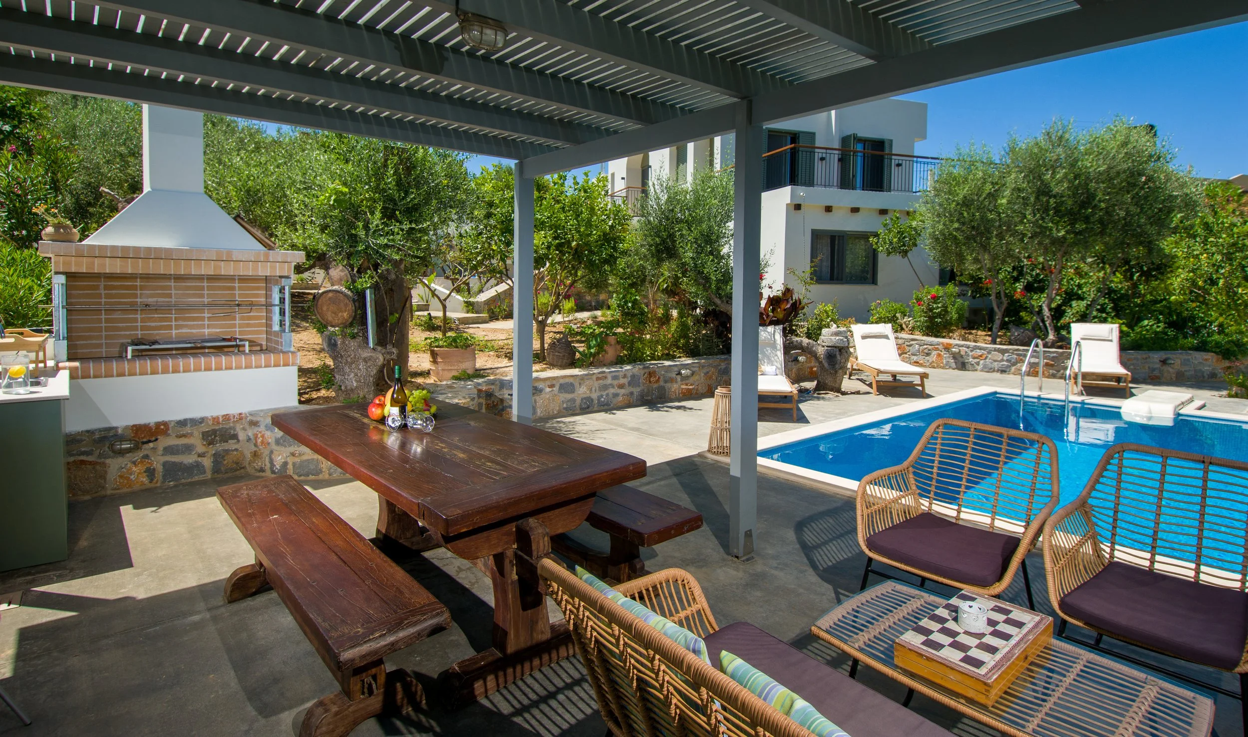 A backyard patio with a swimming pool, lounge chairs, a wooden table with drinks and fruits, a brick outdoor fireplace, and greenery around.