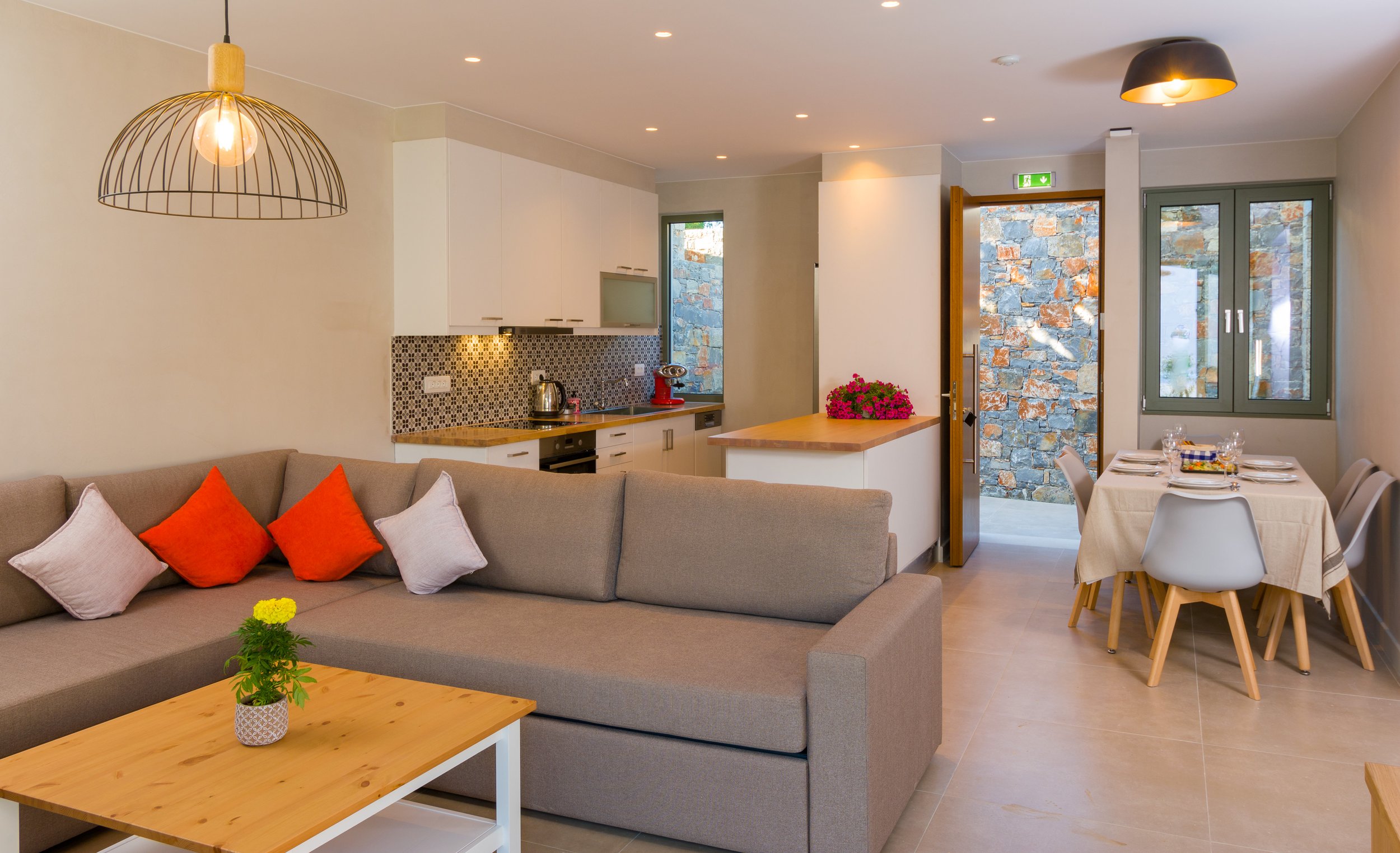 Interior of a modern, open-plan living room and kitchen with a beige sofa, colorful pillows, wooden coffee table with plant, kitchen area with white cabinets, countertops, backsplash, dining table set for four, and large windows and door with stone w