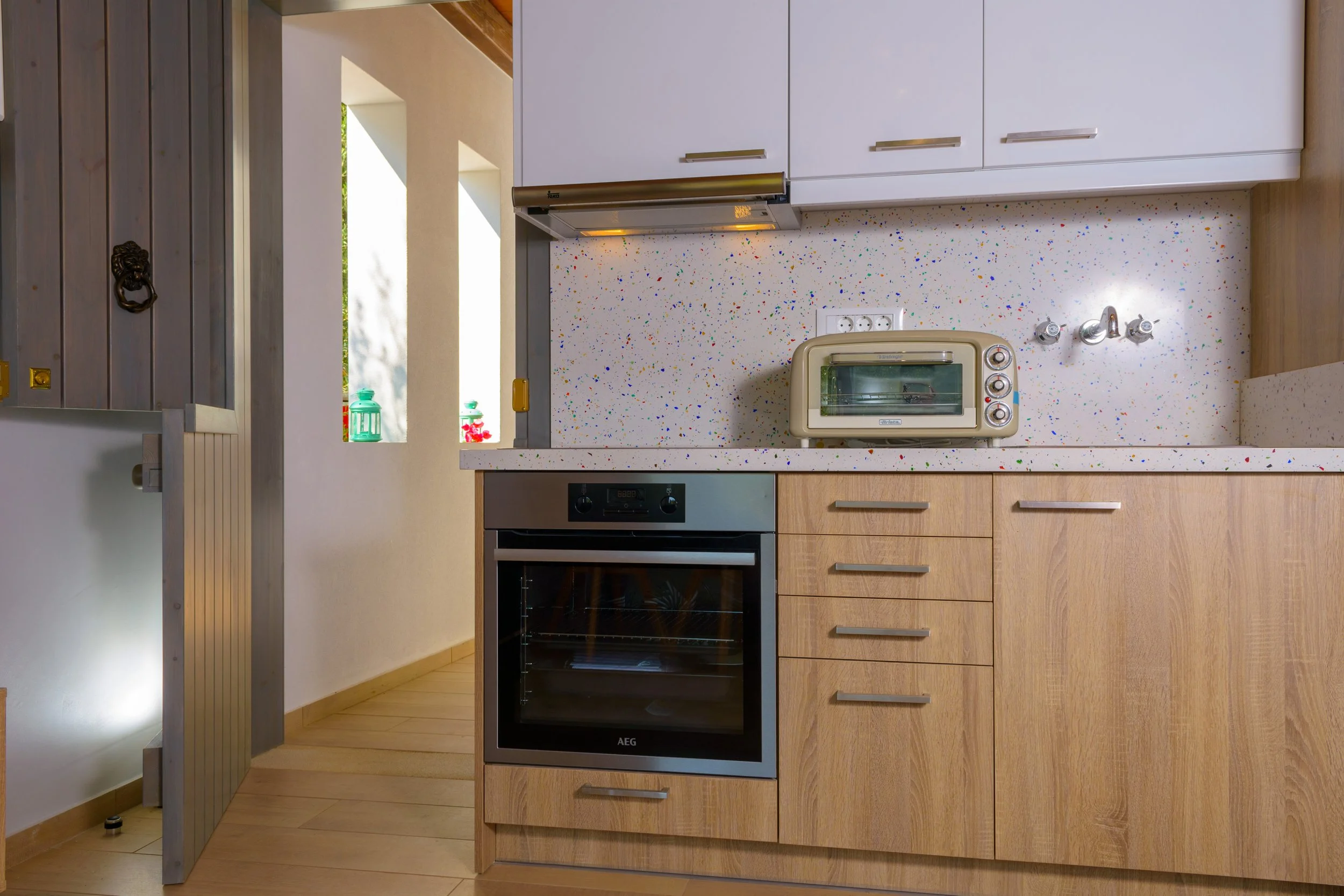 A modern kitchen with wooden cabinetry, a speckled white countertop, a built-in oven, and a small toaster oven on the counter. There are two electrical outlets and a metallic faucet on the wall. Part of a door or partition with a lion head door knock