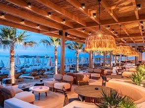 Outdoor seaside lounge area with palm trees, wooden ceiling, hanging lights, and cushioned seating at sunset.