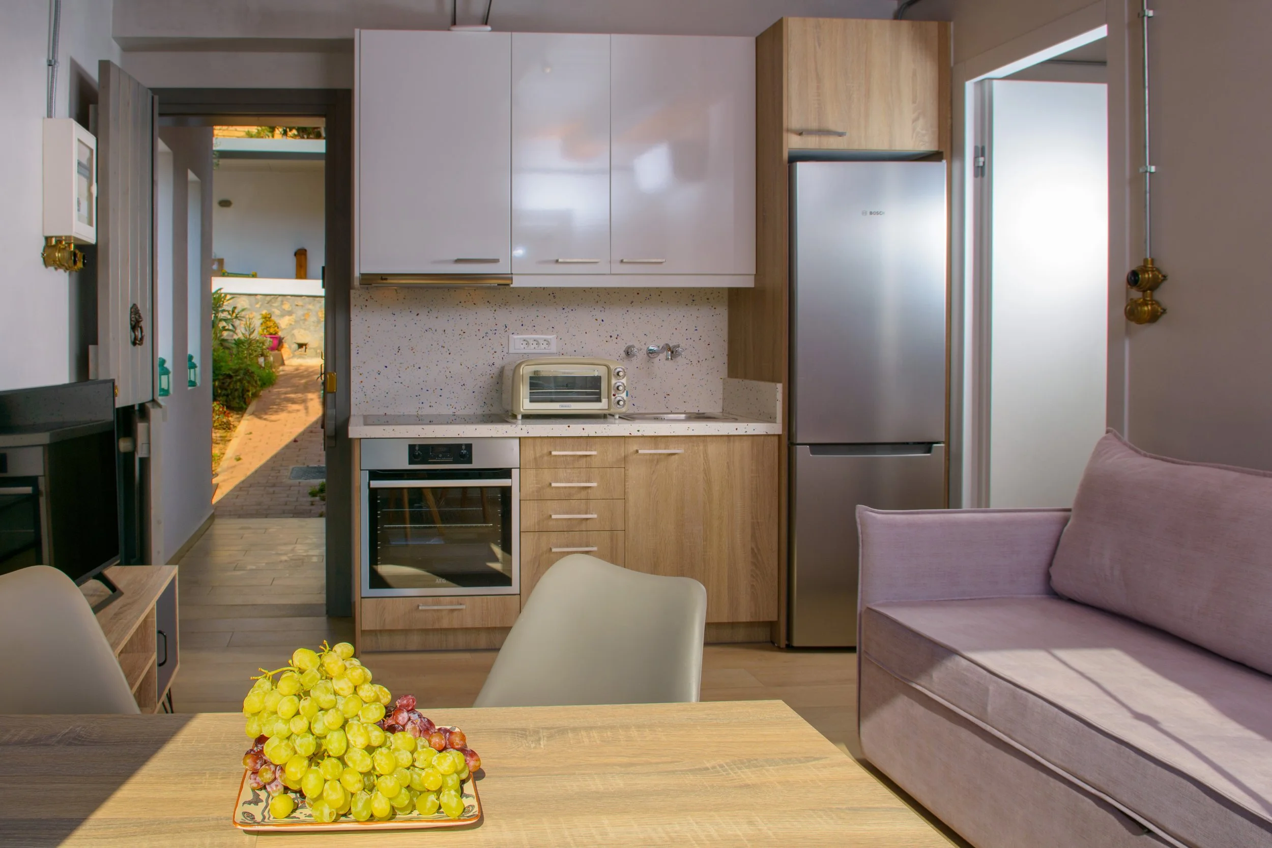A small kitchen with white and wood cabinets, a stainless steel refrigerator, a small oven, and a toaster oven. There is a doorway leading outside and part of a pink sofa and a table with a bunch of grapes and other fruit.