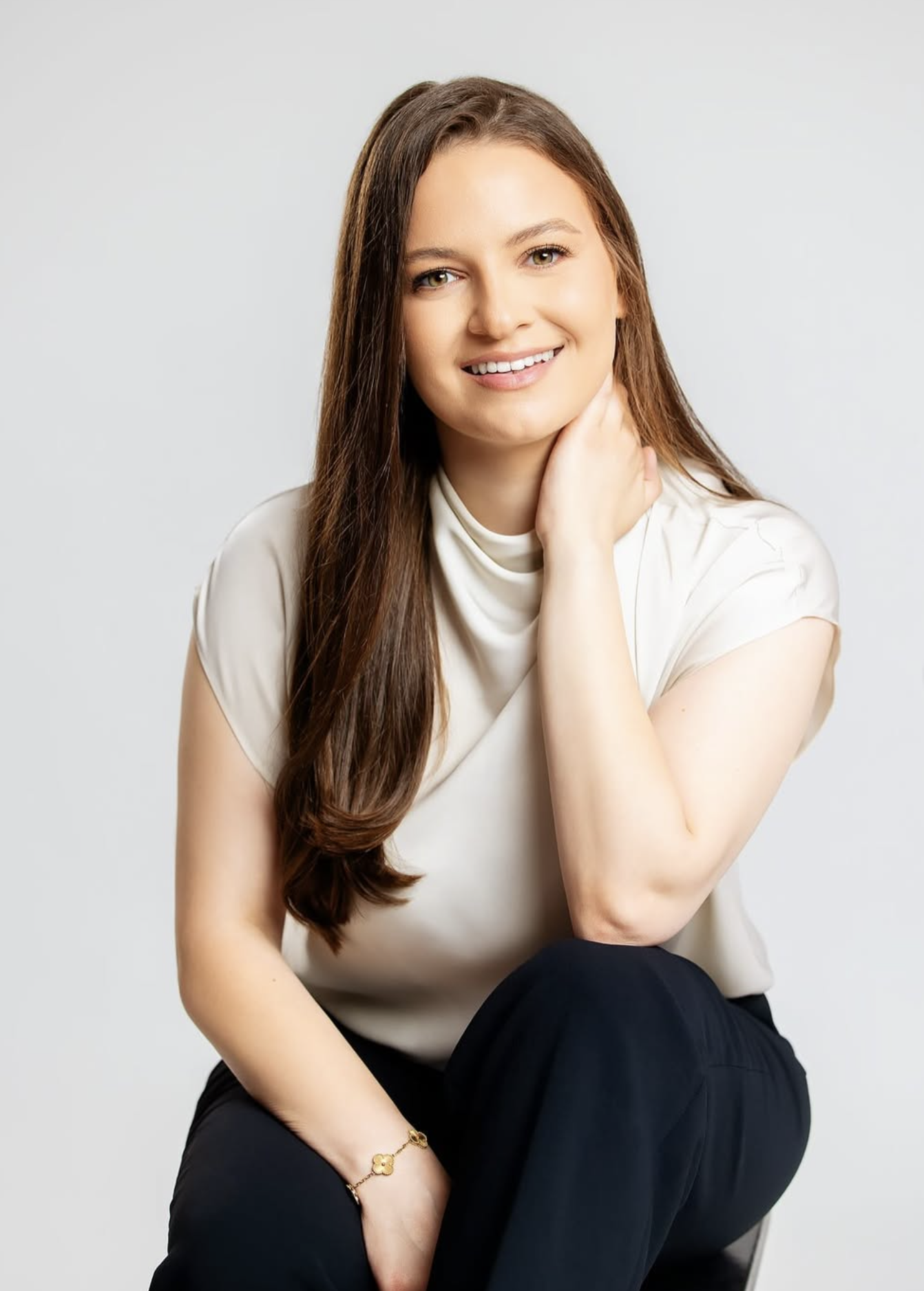 A young woman with long, wavy brown hair, smiling, sitting against a plain, light gray background. She features a fair complexion and is dressed in a light-colored short-sleeve top, black pants, a bracelet, and a ring, with one hand resting on her neck.