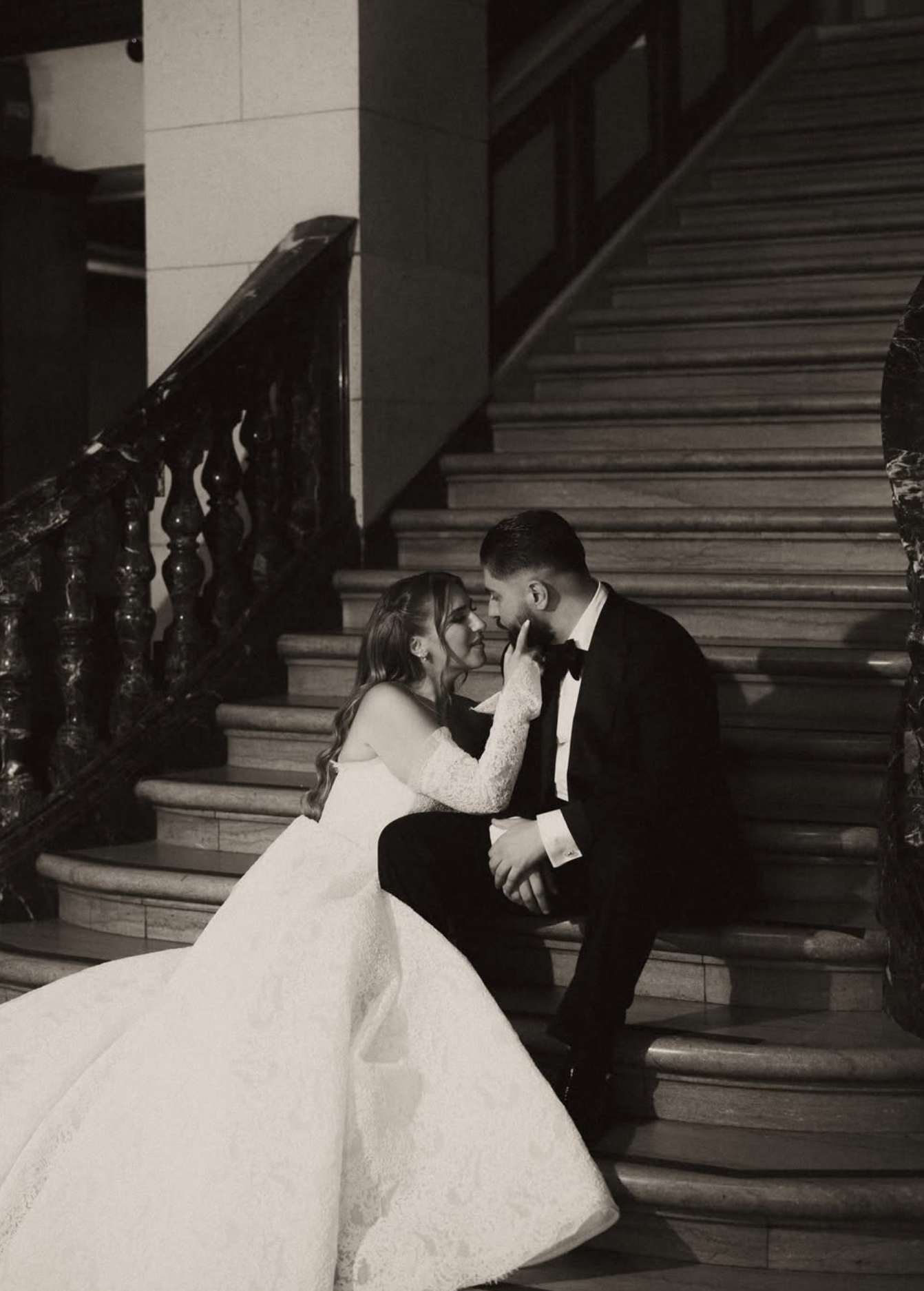 A black and white photo of a bride and groom sitting on the stairs inside a building. The bride is holding the groom's chin gently, and they are gazing into each other's eyes.