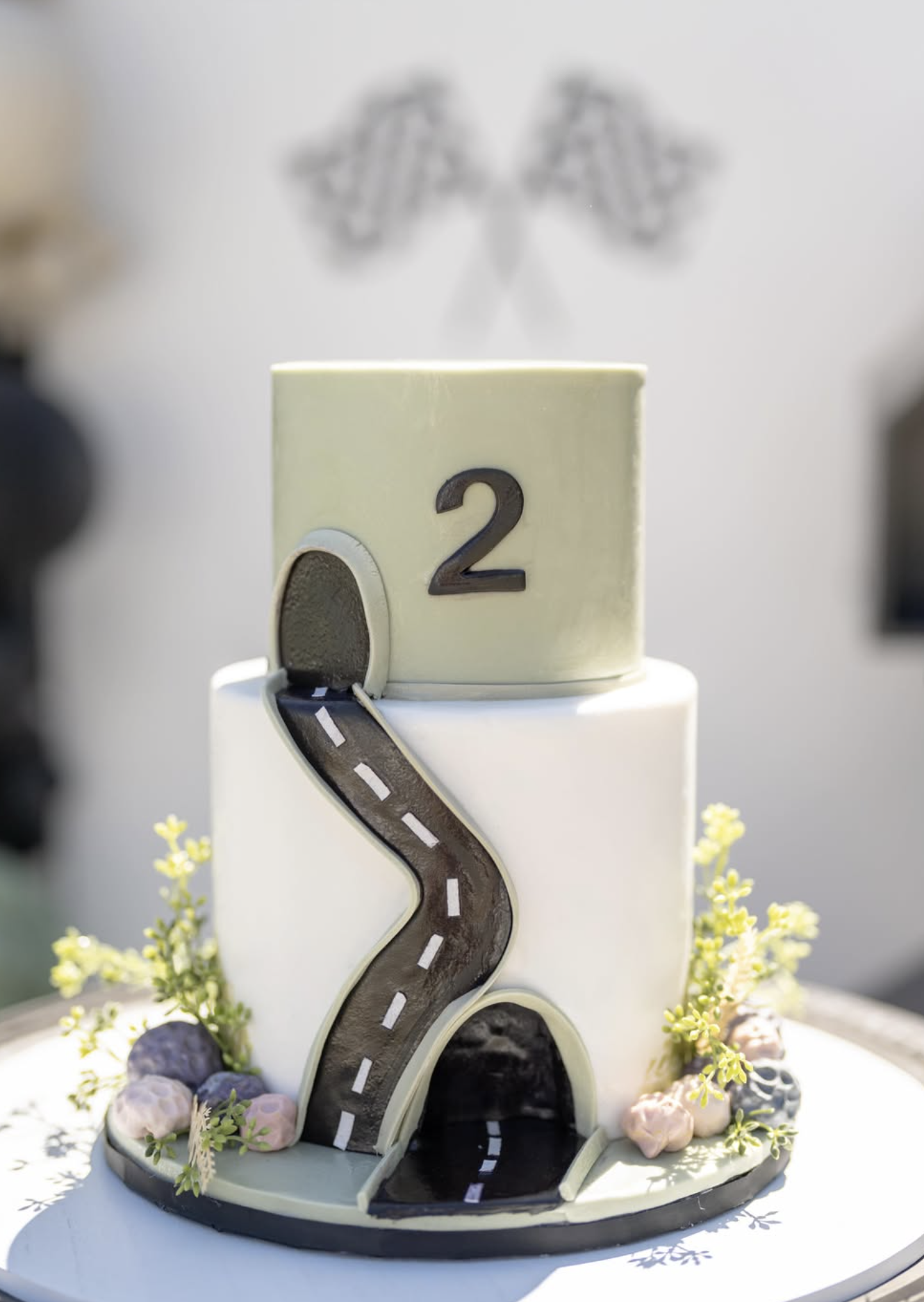 A wedding cake decorated with a black road-shaped topper, with a number 2 on top, surrounded by small flowers and berries.