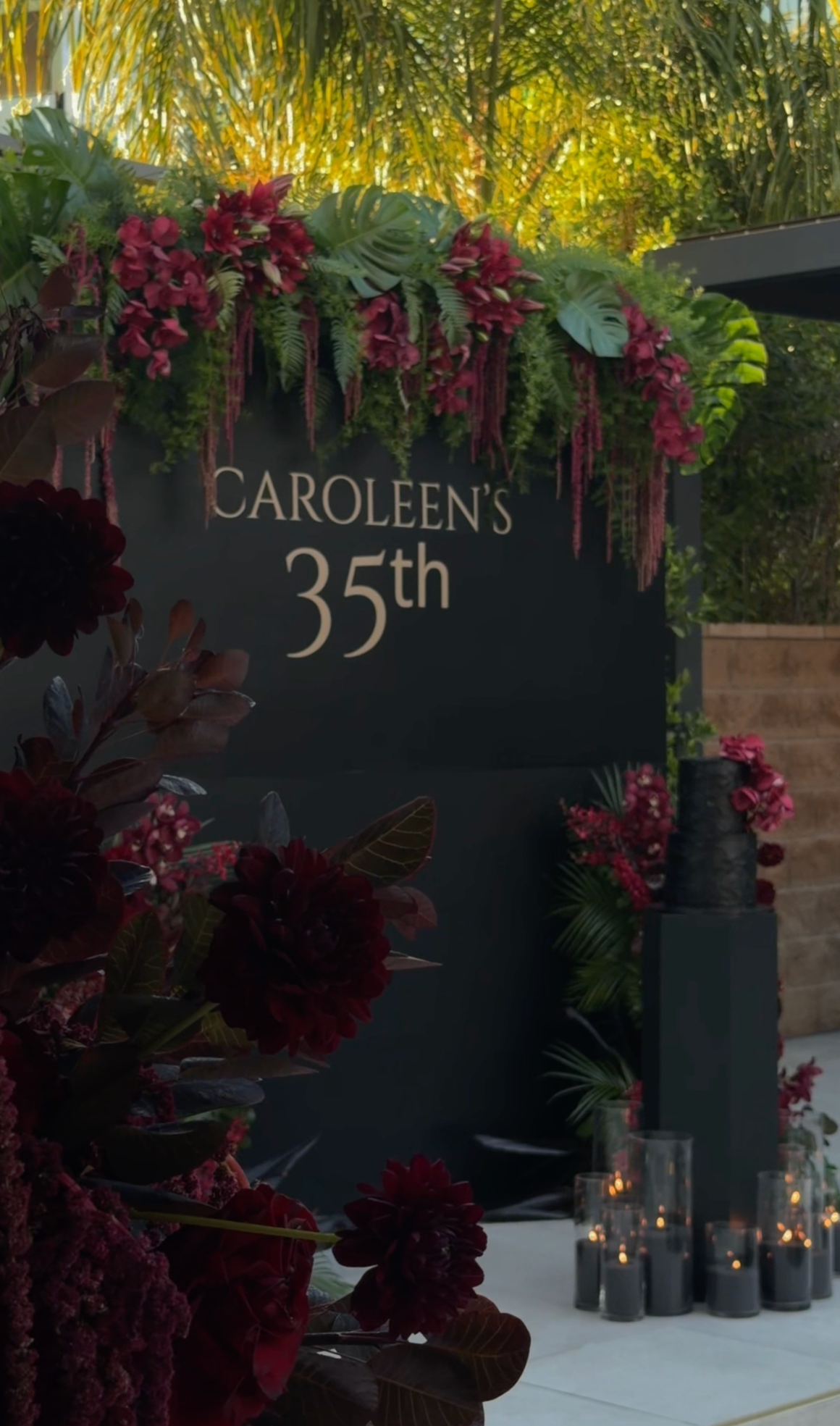 A sign reads 'Caroleen's 35th' surrounded by red and pink floral arrangements and tropical green leaves, with glowing candles in glass holders on a white tablecloth in the foreground, during an outdoor celebration.
