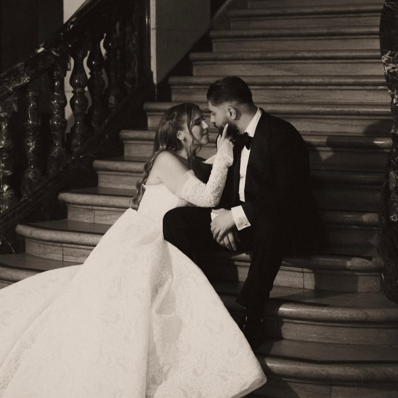 A black and white photo of a bride and groom sitting on a staircase. The bride is wearing a long wedding gown and lace gloves, and the groom is in a black tuxedo. They are gazing into each other's eyes, with the bride touching the groom's face.