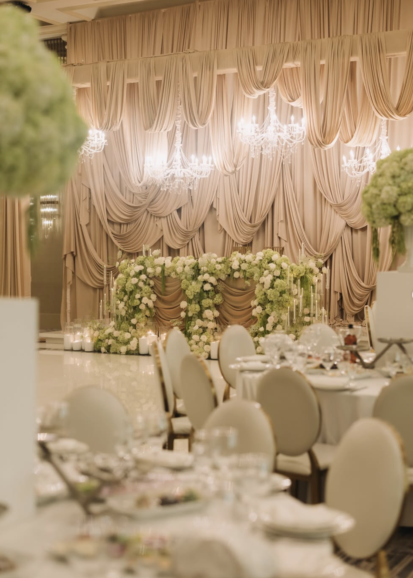 Elegant wedding reception venue with beige draped curtains, chandeliers, and floral arrangements of white and green flowers on the stage.