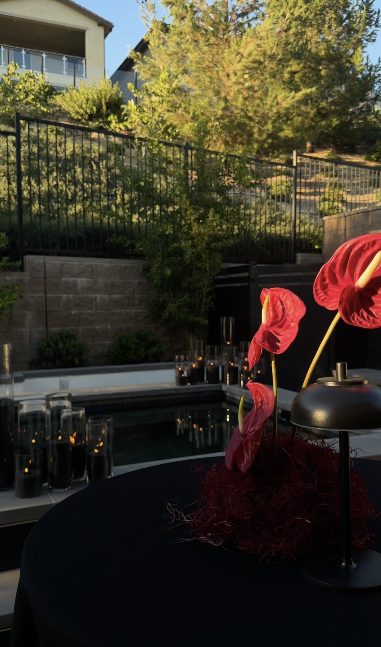 Outdoor patio with pink anthurium flowers in a black vase on a dining table, surrounded by candle holders, overlooking a garden with trees and residential buildings.