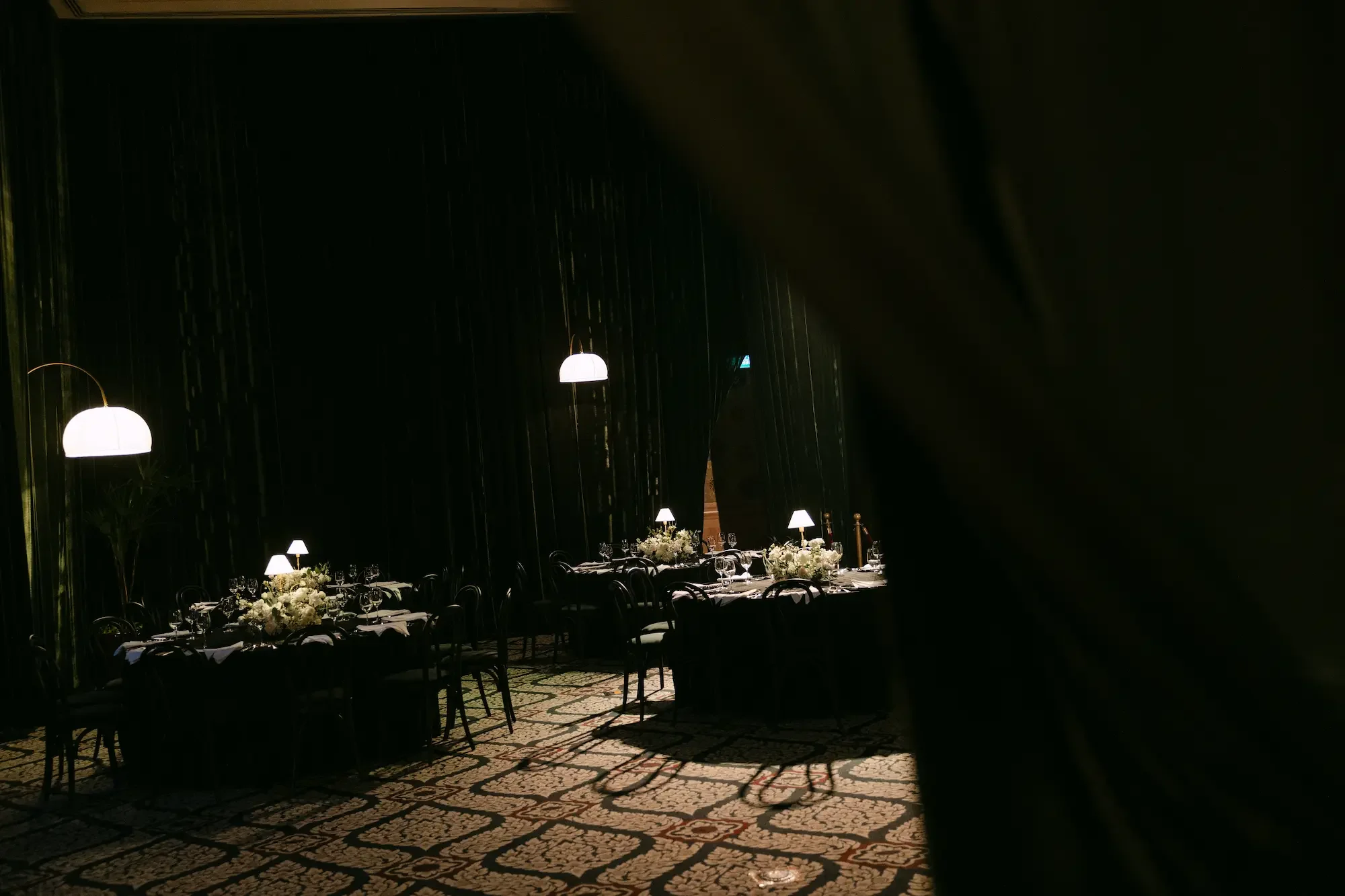 Elegant dining room with long table decorated with white floral arrangements, set with wine glasses and plates. The room has dark walls, hanging pendant lights, and patterned carpet flooring.