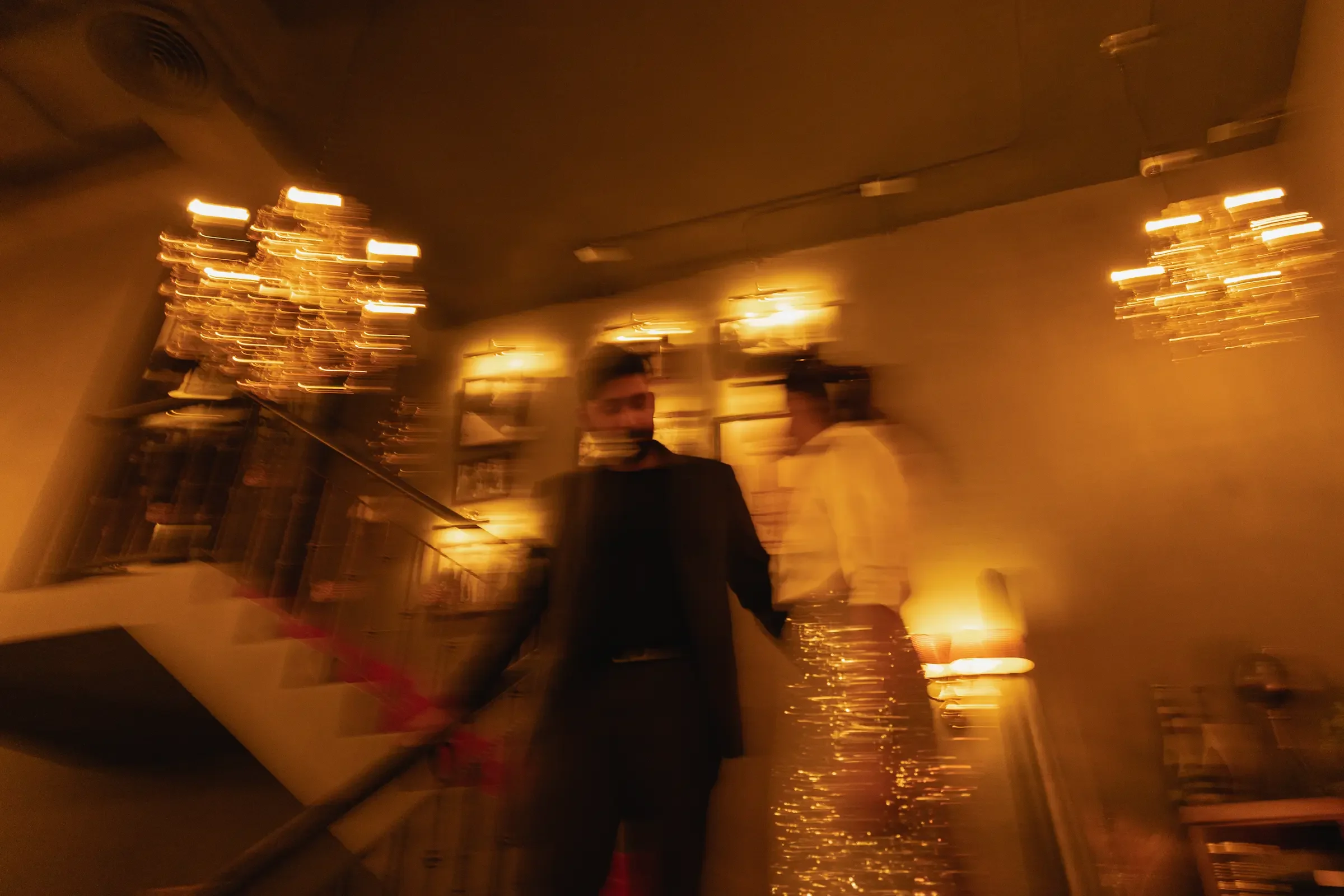 Two guests in a warmly lit wedding reception hall with chandeliers and framed pictures, captured in soft motion blur.