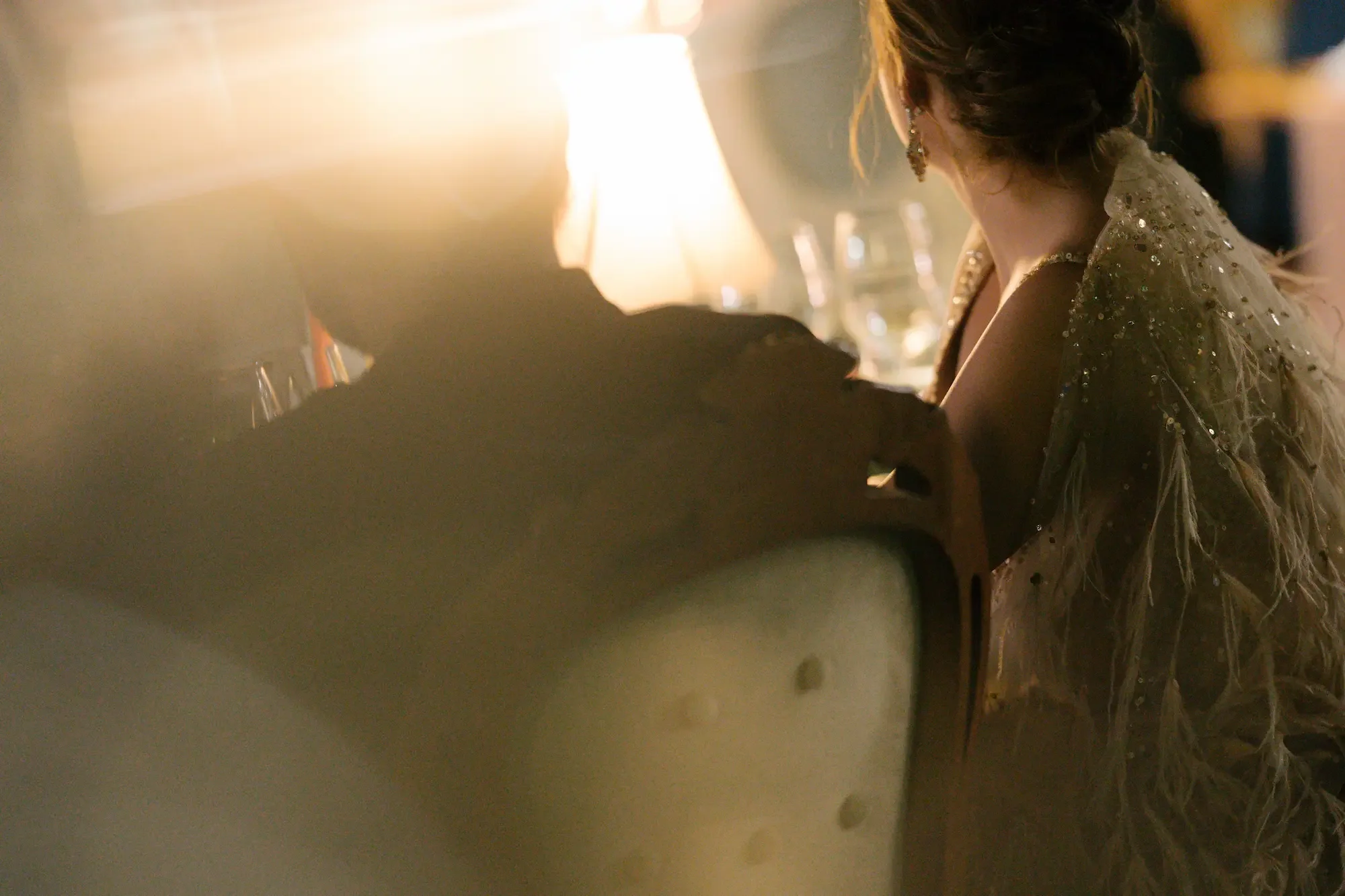 A woman dressed in a decorated gown with beads or sequins, and earrings, facing away with her head turned to the side, showing her profile, in a setting with warm, soft lighting.