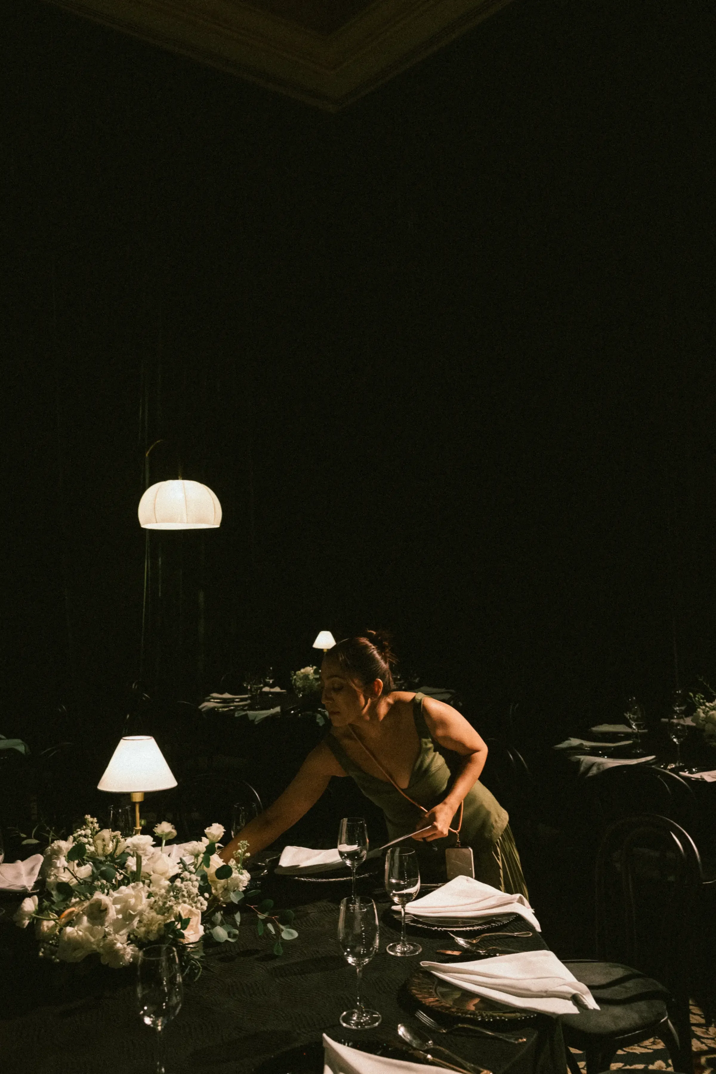 A woman arranging a floral centerpiece at a formal dining table in a dimly lit restaurant with hanging pendant lights and elegantly set tables.