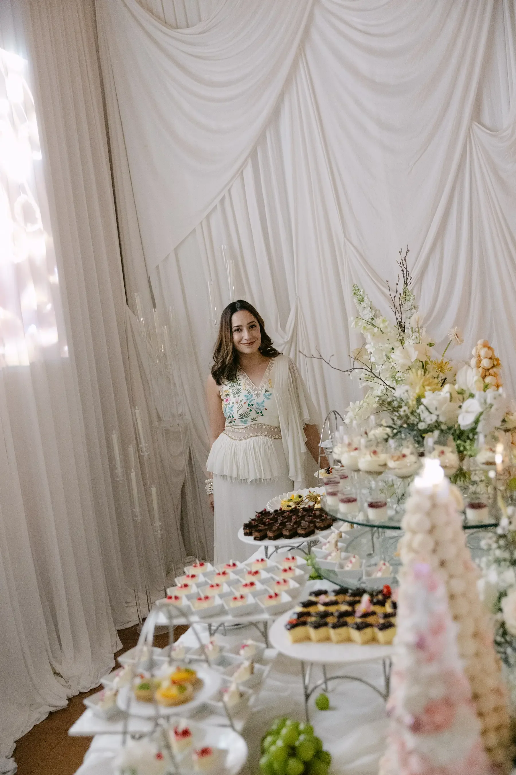 A woman in a white dress standing next to a table filled with various desserts and a floral arrangement. The background features white draped curtains. The desserts include small cakes, fruit slices, and other sweet treats displayed on tiered trays.