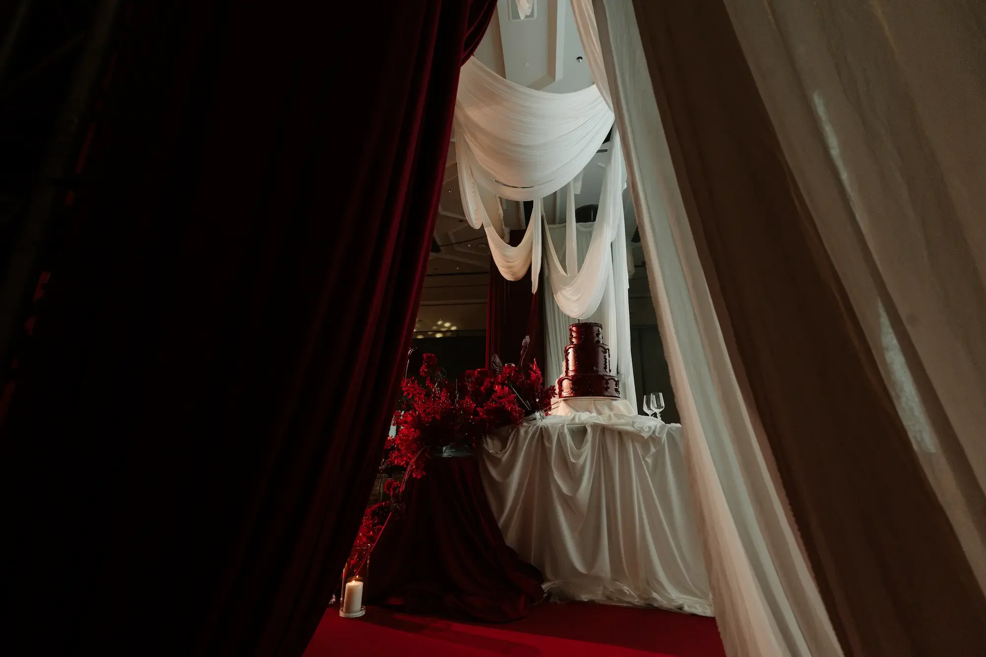 A wedding reception table decorated with white drapery, a wedding cake, red flowers, and candles, viewed through a partially open red curtain.