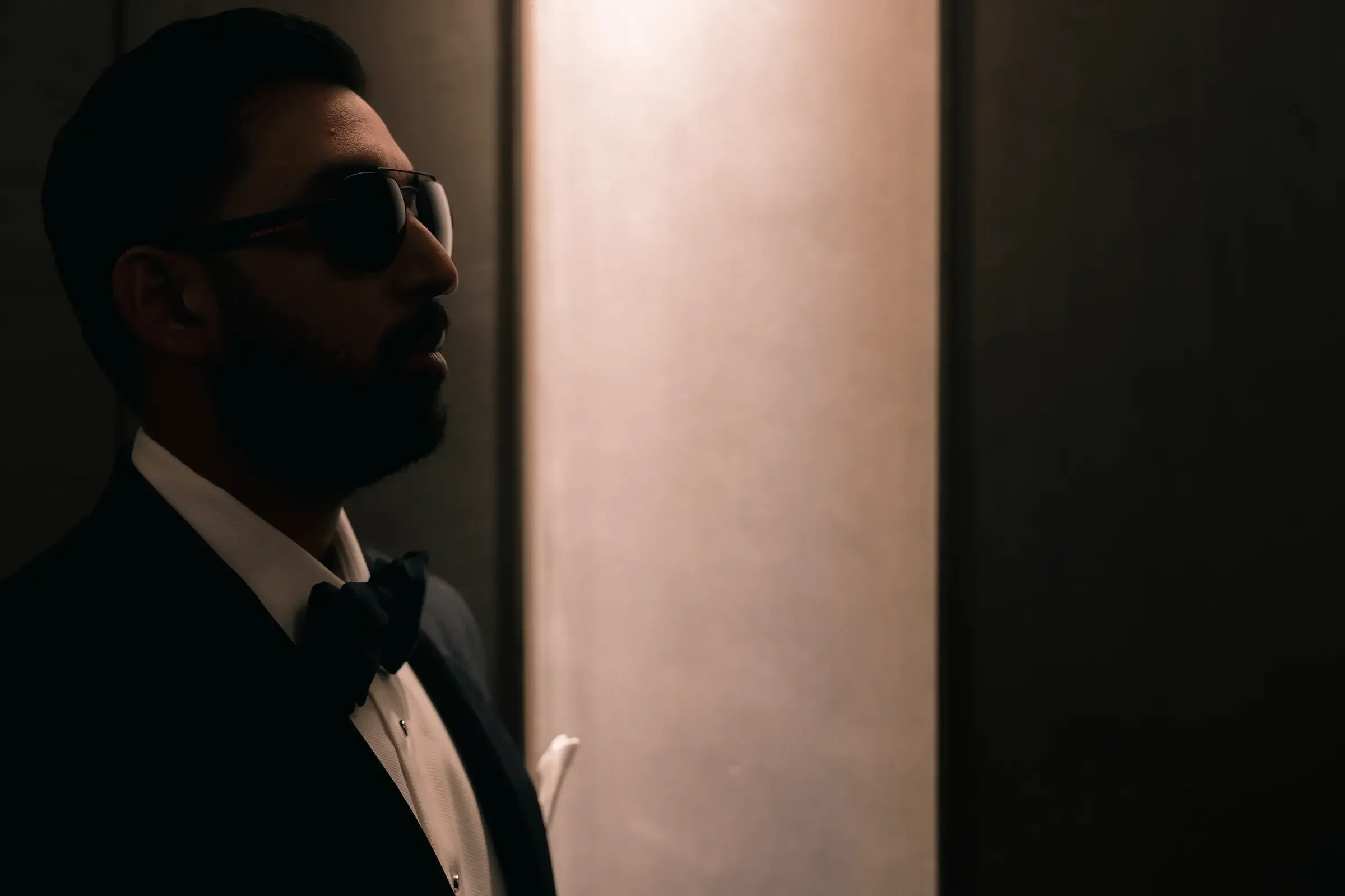 A man wearing sunglasses, a tuxedo, and a bow tie, standing in an elevator with a plain wall background.