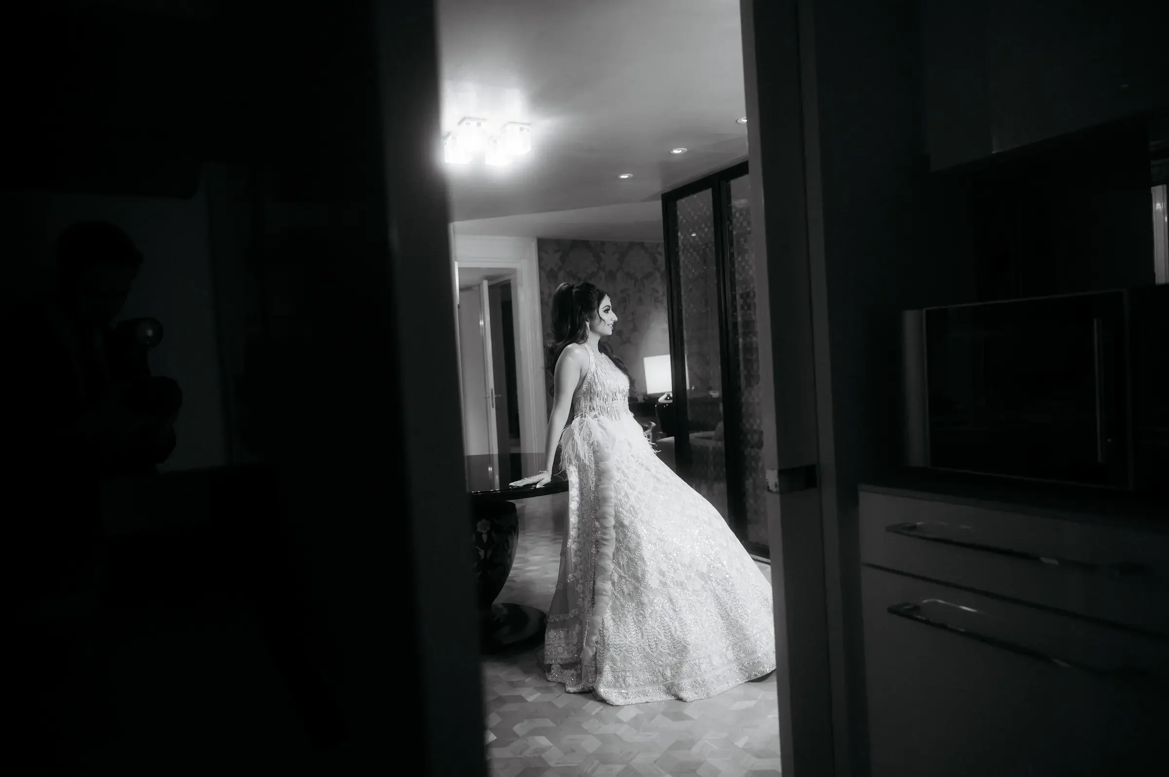 Black and white photo of a woman in a wedding dress, sitting and leaning on a piano, in a modern interior with patterned wall and a lamp in the background.