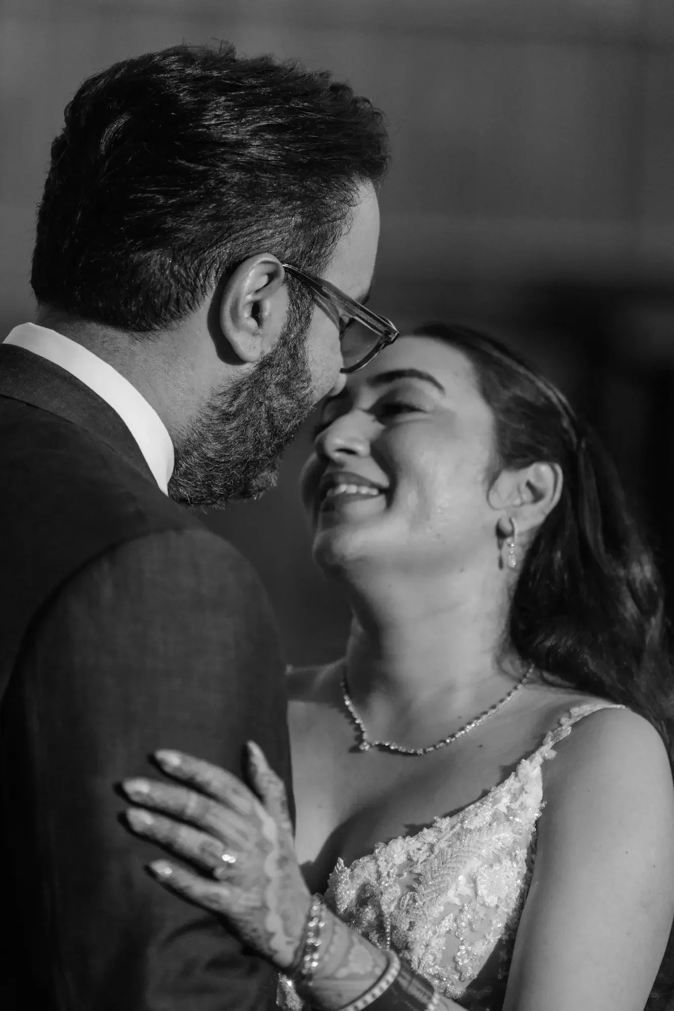 A black and white photo of a man and woman dancing closely. The man has dark hair, glasses, and a beard, while the woman has dark hair, earrings, and a necklace. They are smiling and appear to be sharing an intimate moment.
