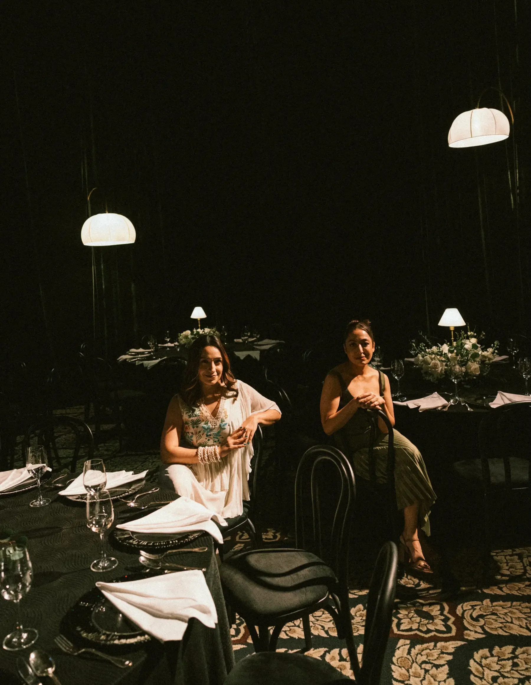 A dimly lit restaurant with a dark ceiling and elegant hanging light fixtures. Two women are sitting at a table with white napkins, wine glasses, and floral centerpieces, smiling at the camera.